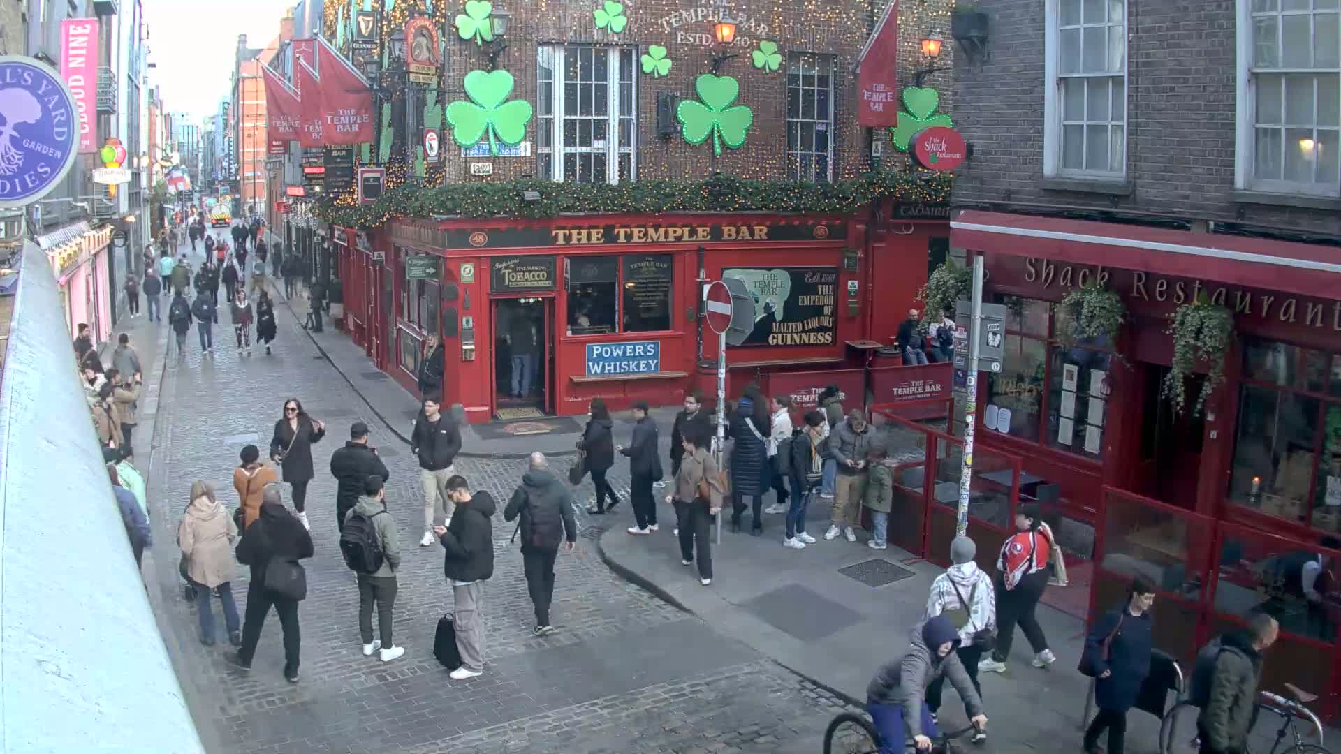 A bustling cobblestone street scene shows many pedestrians walking past red brick buildings, one of which is a pub decorated with shamrocks and holiday lights, on an overcast day.