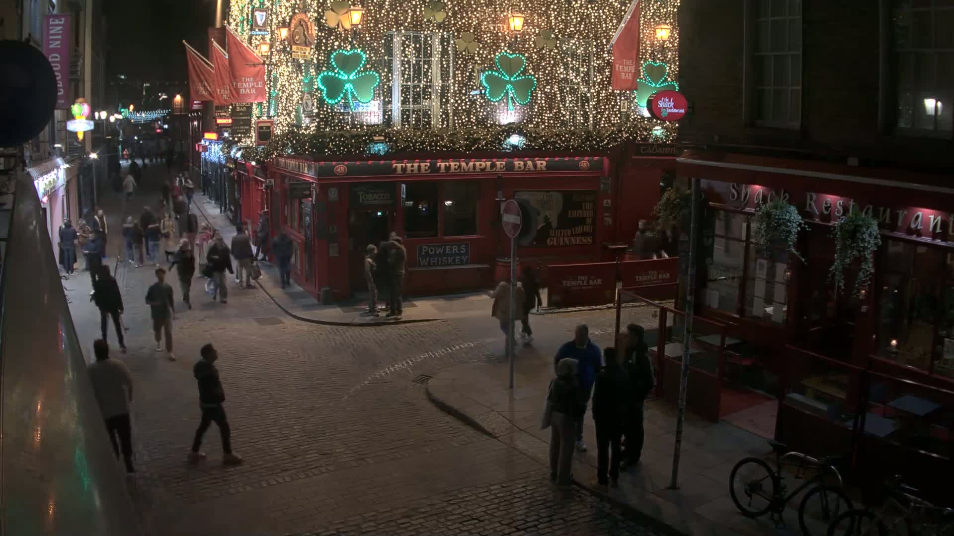 At night, a cobblestone street is bustling with people near a brightly lit, festively decorated pub.