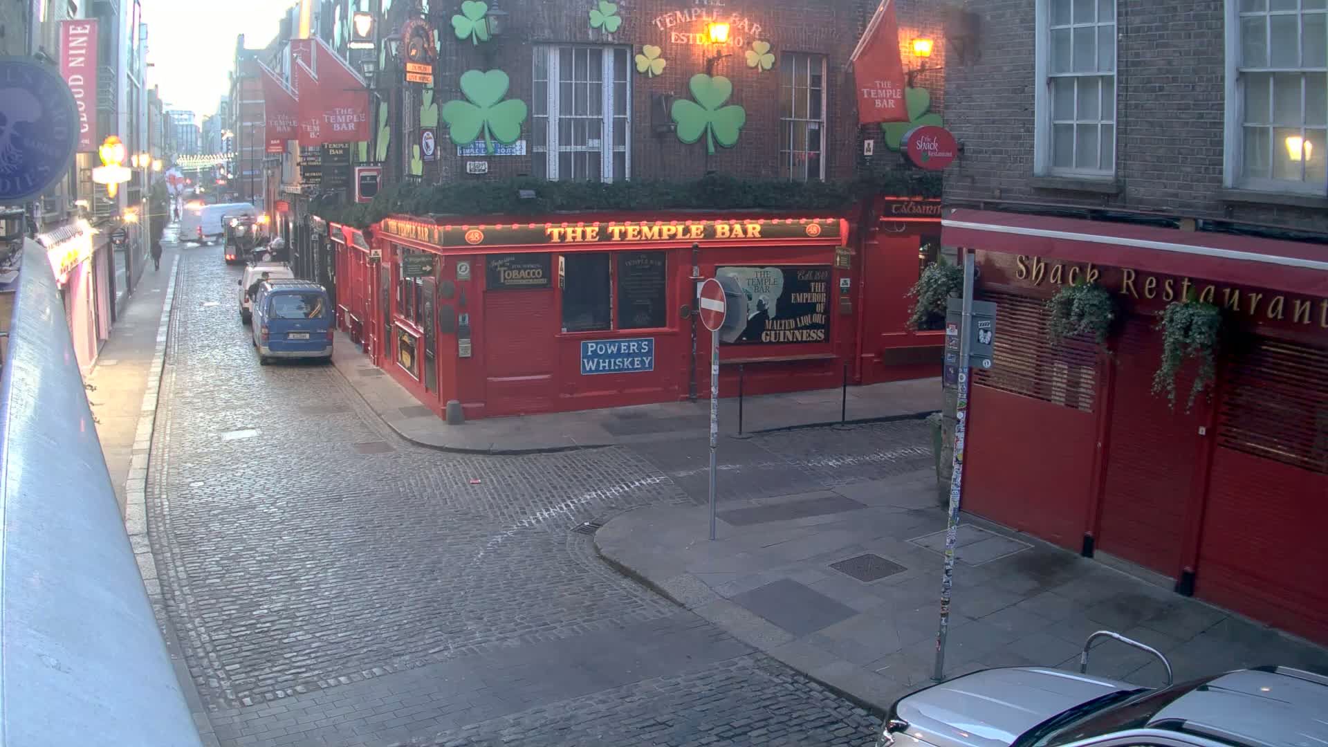 A cobblestone street scene at dusk shows a red building called "The Temple Bar" with several vehicles parked nearby.