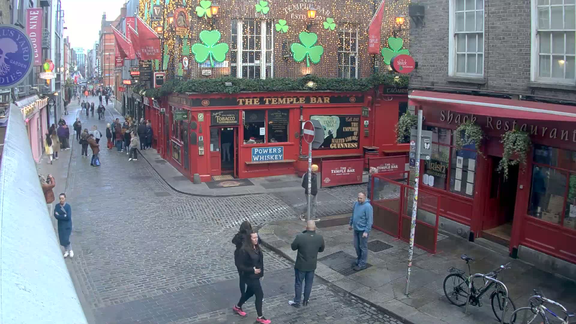 A cobblestone street, lined with red brick buildings decorated with shamrocks and string lights, shows several pedestrians and a bicycle, under an overcast sky.