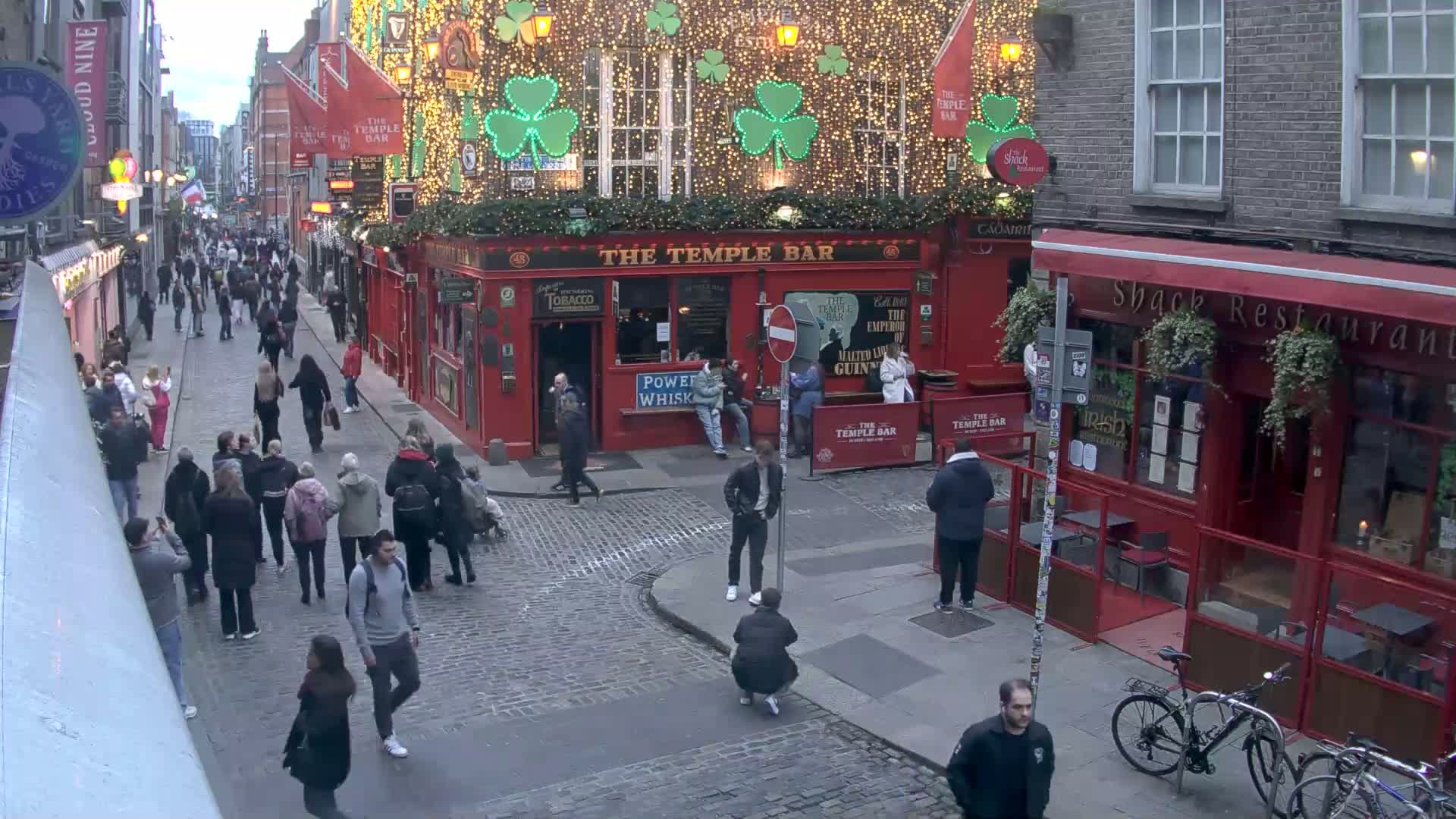 A bustling cobblestone street scene at dusk, with pedestrians walking past a red-brick building adorned with shamrock lights and various businesses, under an overcast sky.