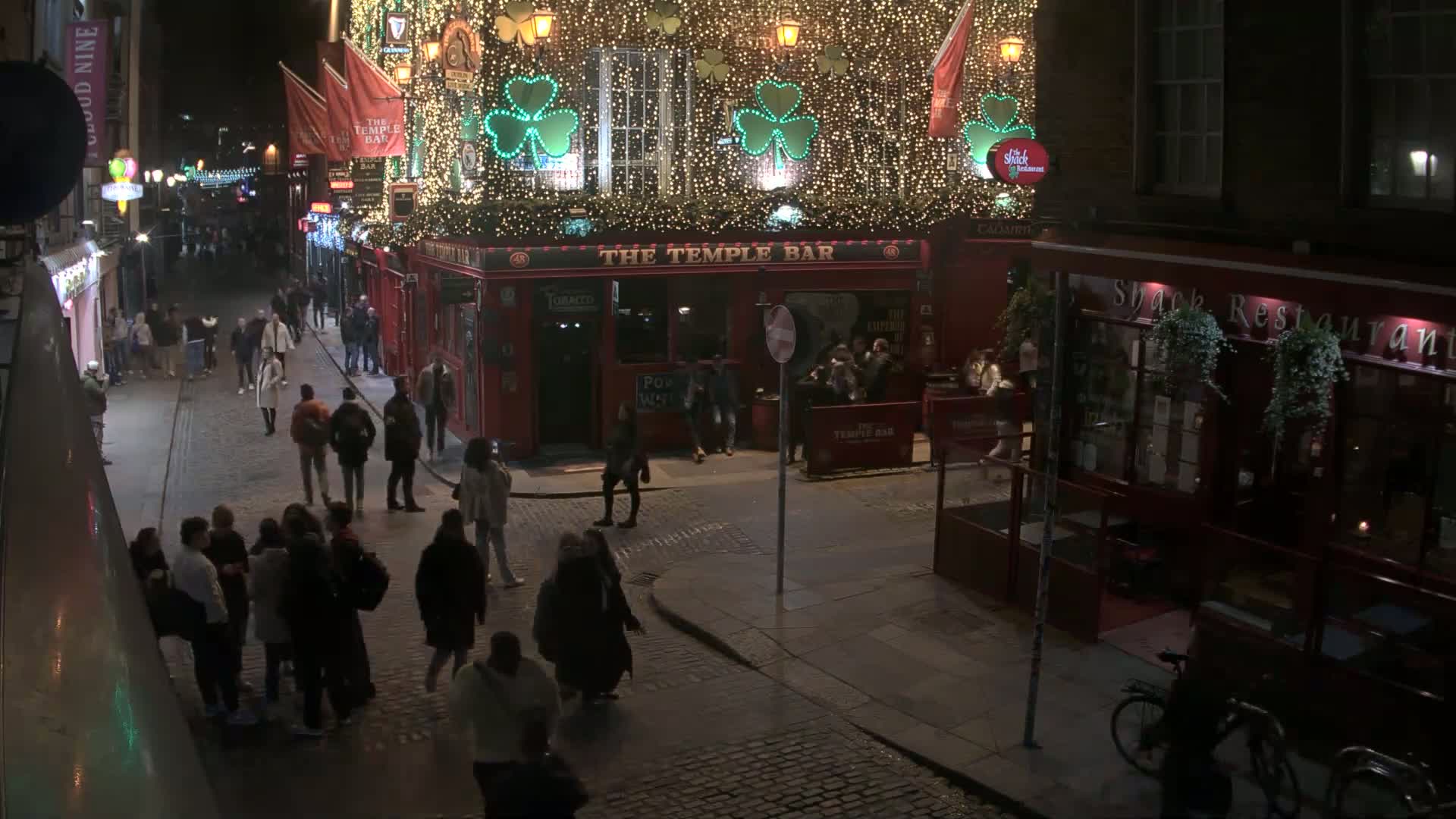 A nighttime street scene shows a cobblestone street with pedestrians and buildings, one of which is brightly lit and decorated with shamrocks and lights.