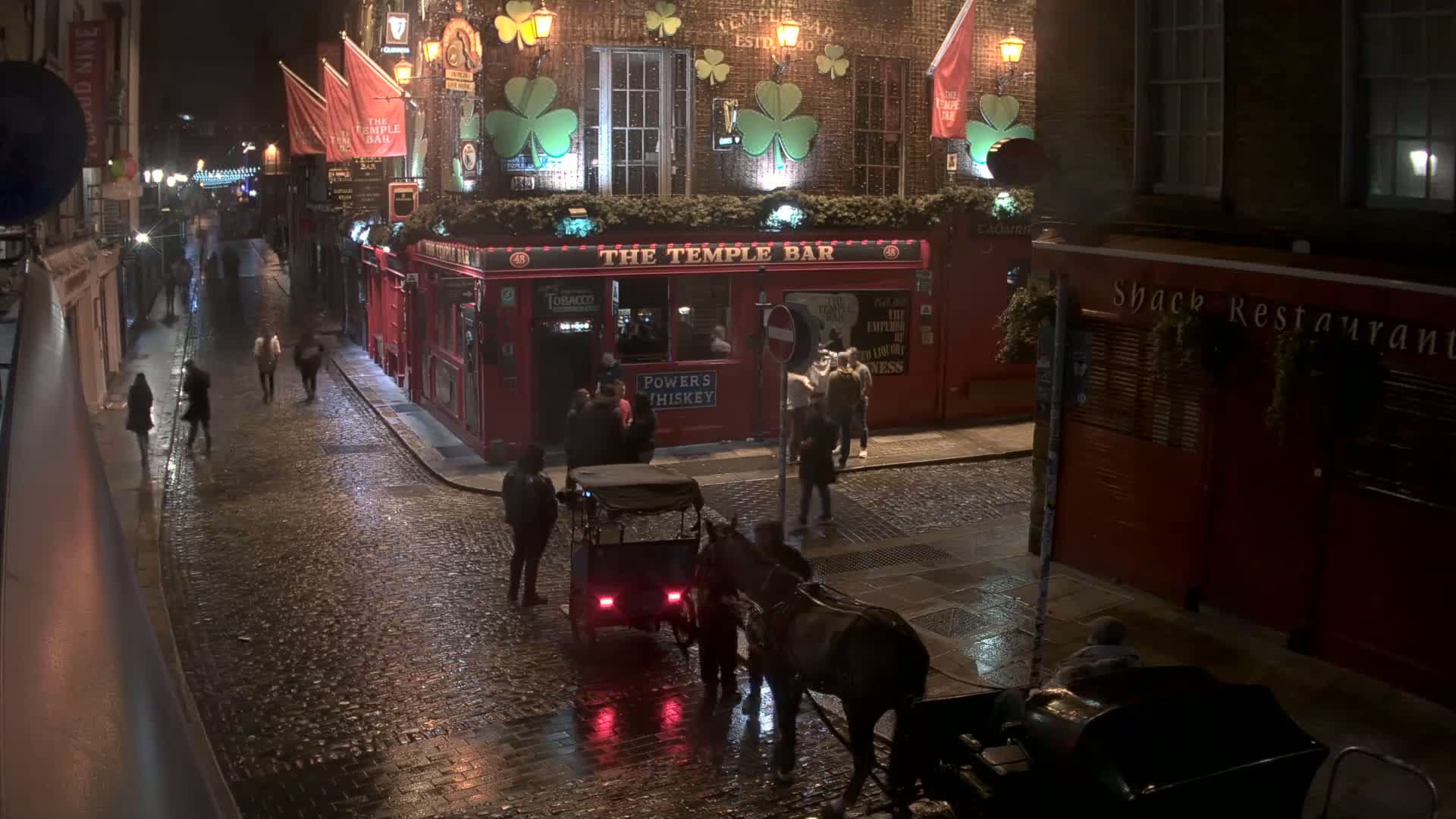 A wet cobblestone street at night shows a horse-drawn carriage and several people walking past a red brick pub.