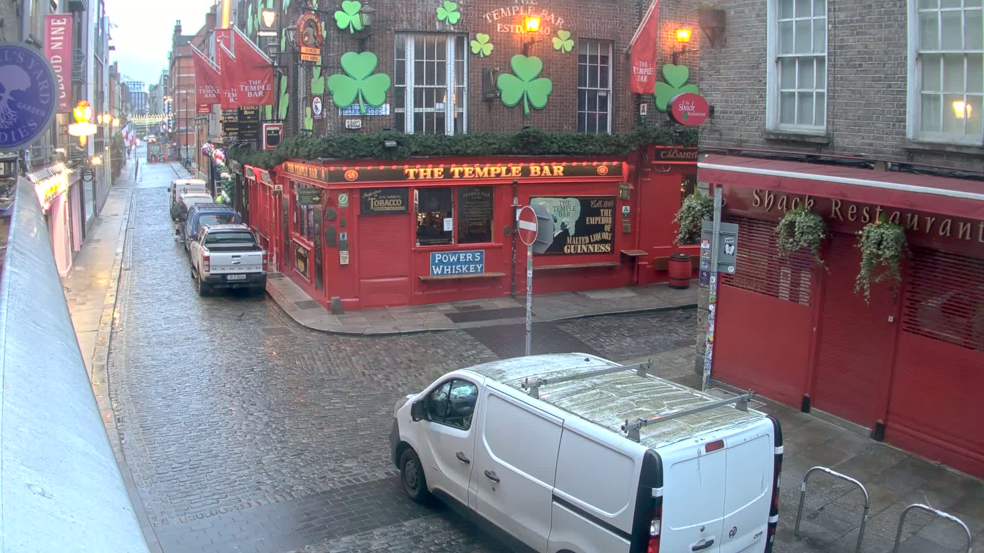 A wet cobblestone street, on a cloudy day, shows a red brick building with a pub called "The Temple Bar," parked vehicles, and a white van.
