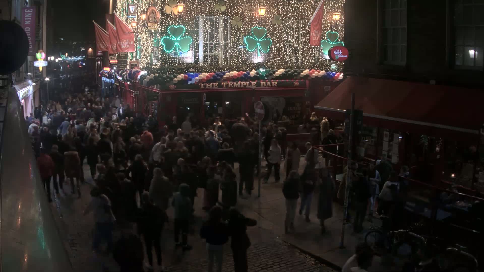 A large crowd of people gathers on a cobblestone street at night outside a brightly lit bar decorated with shamrocks and string lights.