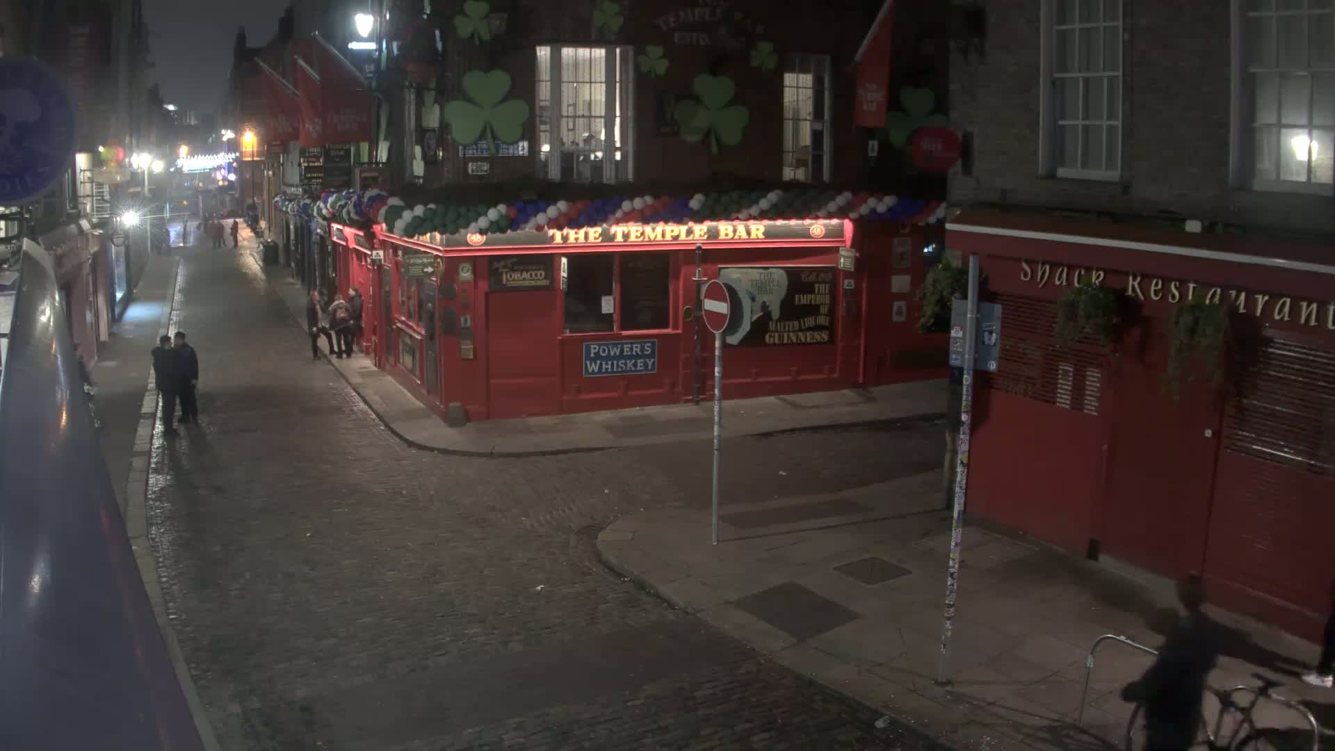A nighttime street scene shows a cobblestone road with several people standing near a red brick building, which is decorated with balloons and has a pub sign.