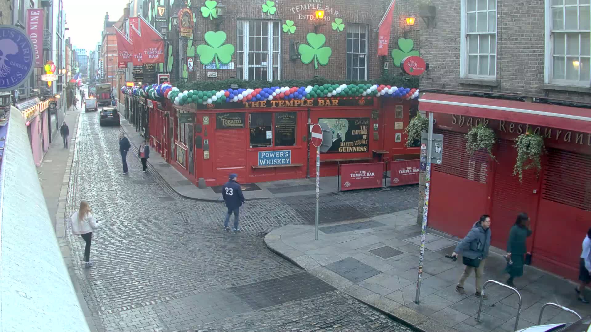 A cobblestone street scene shows pedestrians walking past a red building adorned with shamrocks and balloons, under an overcast sky.