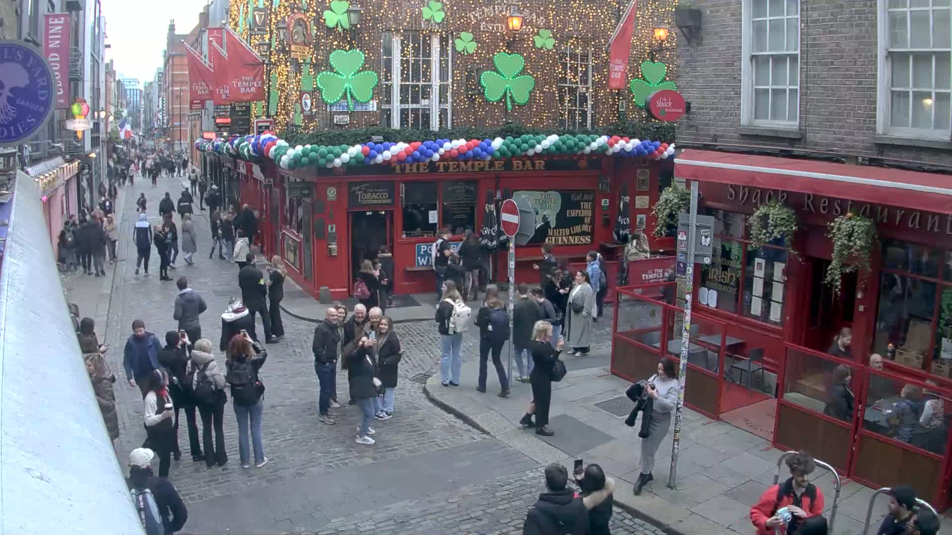 A cobblestone street in a city, possibly Dublin, is filled with pedestrians, and the Temple Bar pub is decorated with shamrocks and balloons on an overcast day.