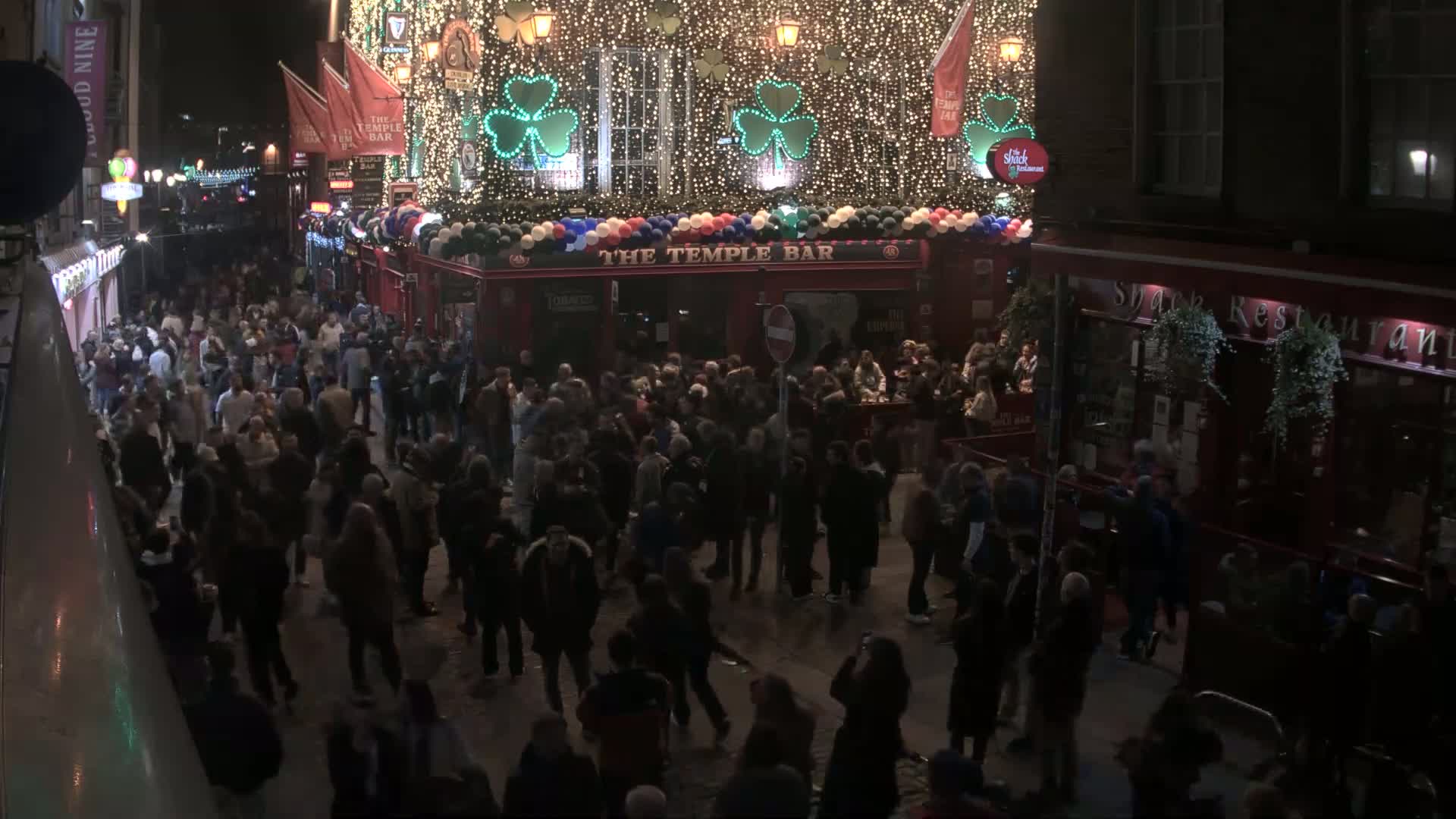 A large crowd of people gathers on a city street at night outside a brightly lit pub decorated with shamrocks and balloons.