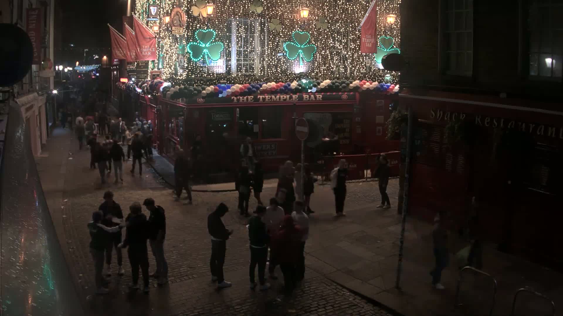 A nighttime street scene shows a crowd of people gathered near a brightly lit bar adorned with shamrocks and string lights, under a dark, clear sky.