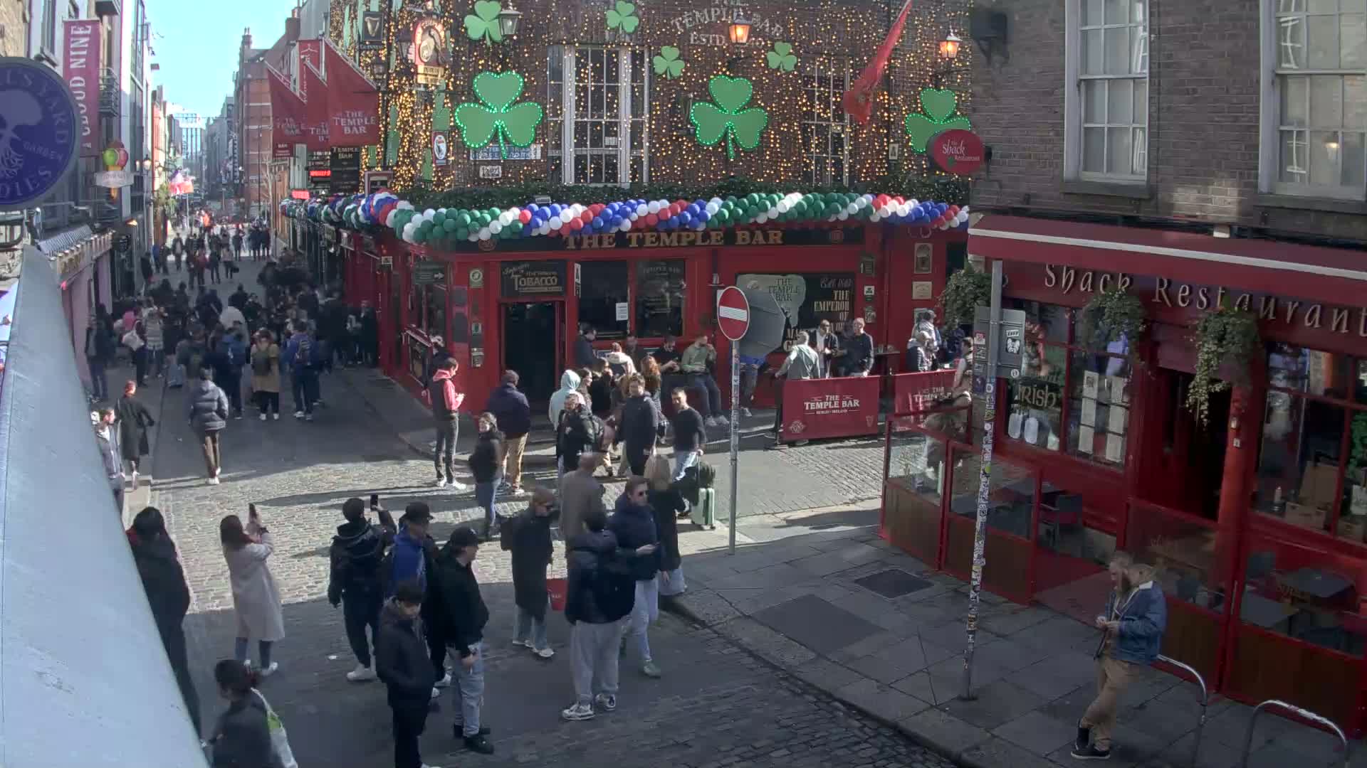 A sunny day reveals a bustling cobblestone street filled with pedestrians, many congregating near a red-brick building decorated with shamrocks and balloons.