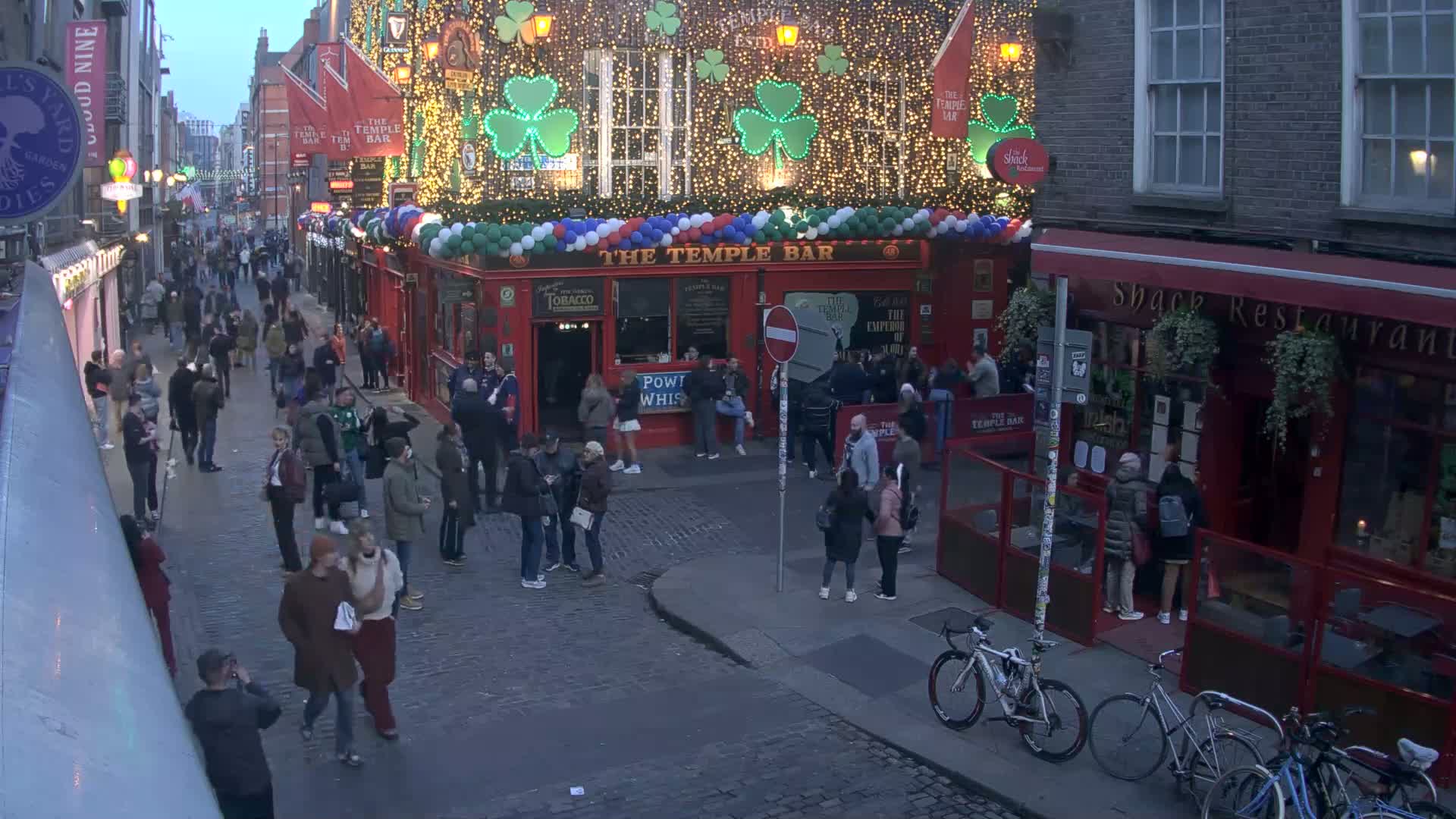 A bustling, cobblestone street scene at twilight, featuring a crowded pub with festive lights and shamrock decorations, and several pedestrians, some bicycles, and other buildings.