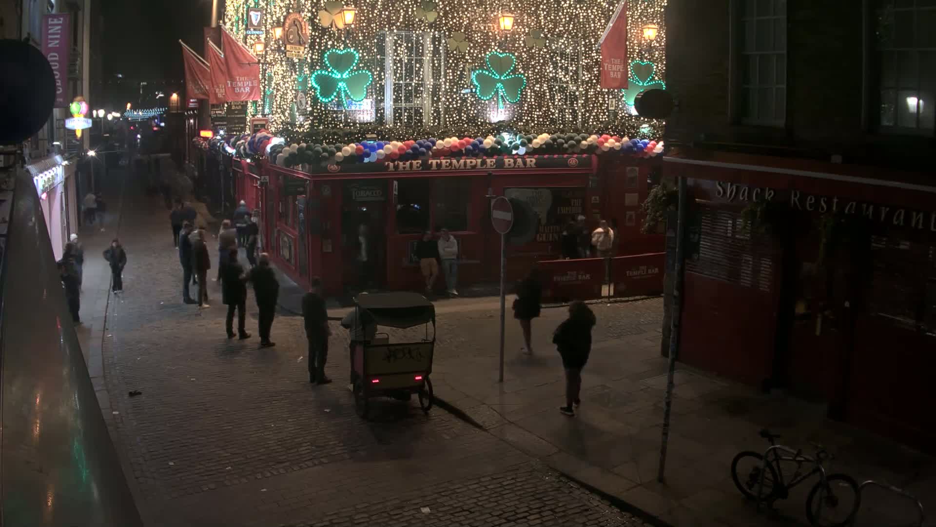 A nighttime street scene shows a cobblestone street with people walking past a brightly lit pub decorated with shamrocks and string lights.