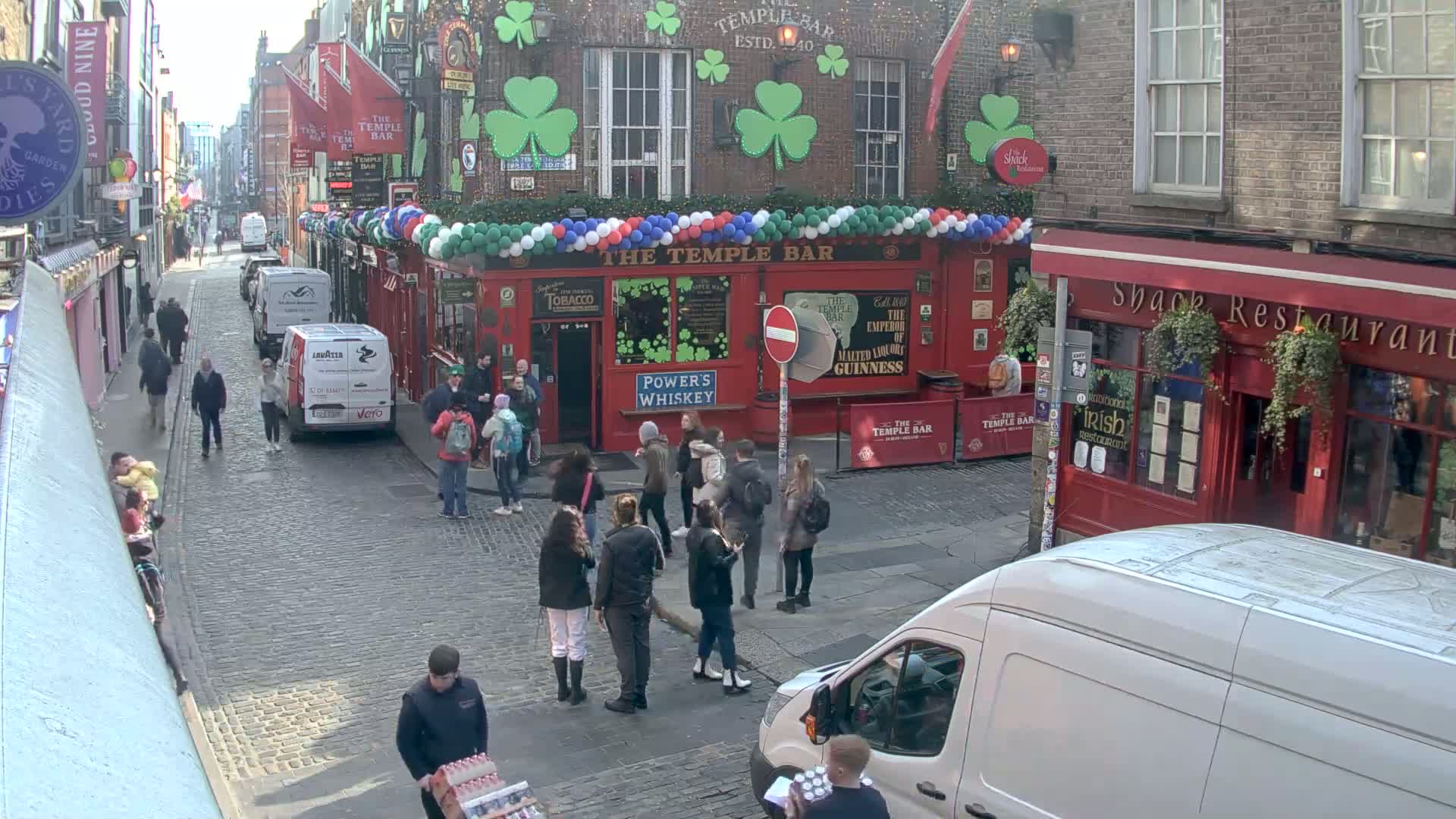 A cobblestone street, bustling with pedestrians, features a red brick pub decorated with shamrocks and balloons, under a sunny sky.