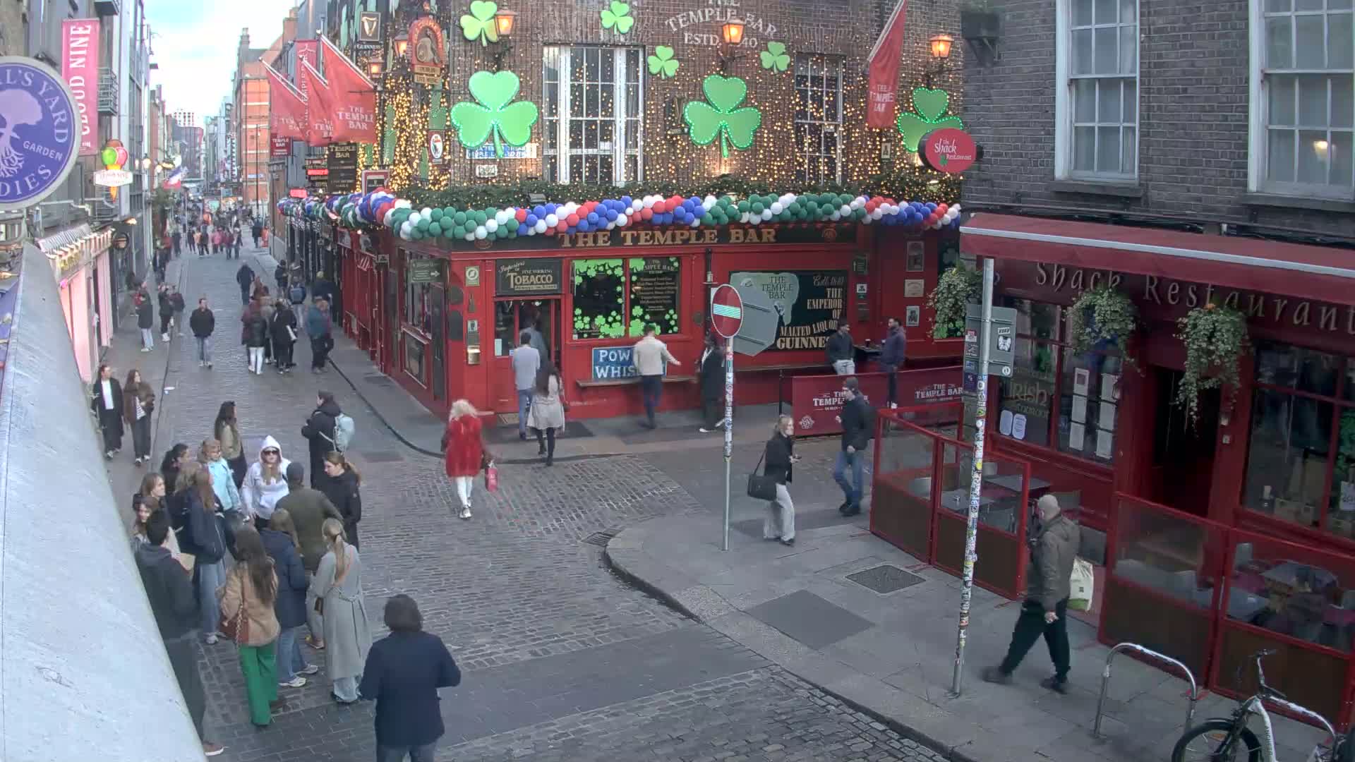 A cobblestone street scene shows pedestrians walking past pubs and restaurants, decorated with shamrocks and balloons, under overcast skies.
