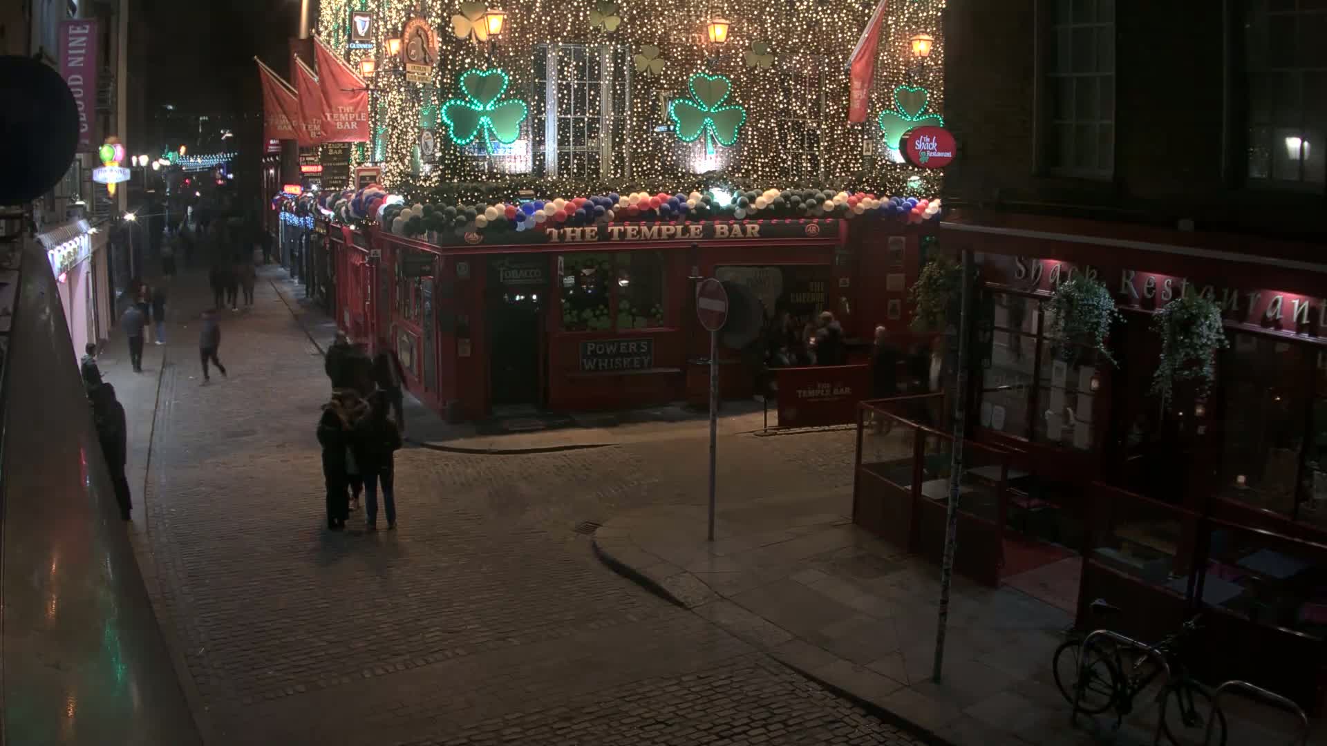 A nighttime street scene shows a cobblestone street with pedestrians and a brightly lit pub decorated with shamrocks and string lights.