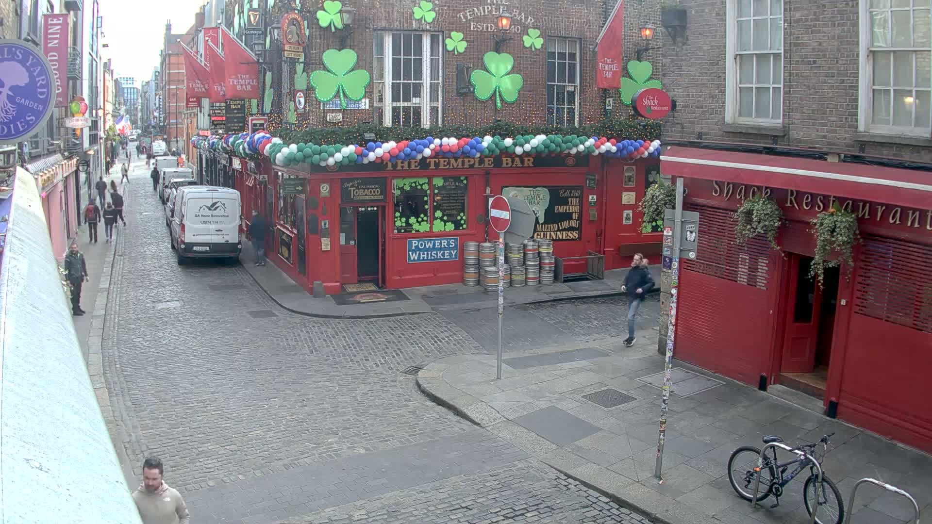 A cobblestone street scene shows a red brick building with a pub called "The Temple Bar," decorated with shamrocks and balloons, next to a red restaurant, with several people walking around on an overcast day.