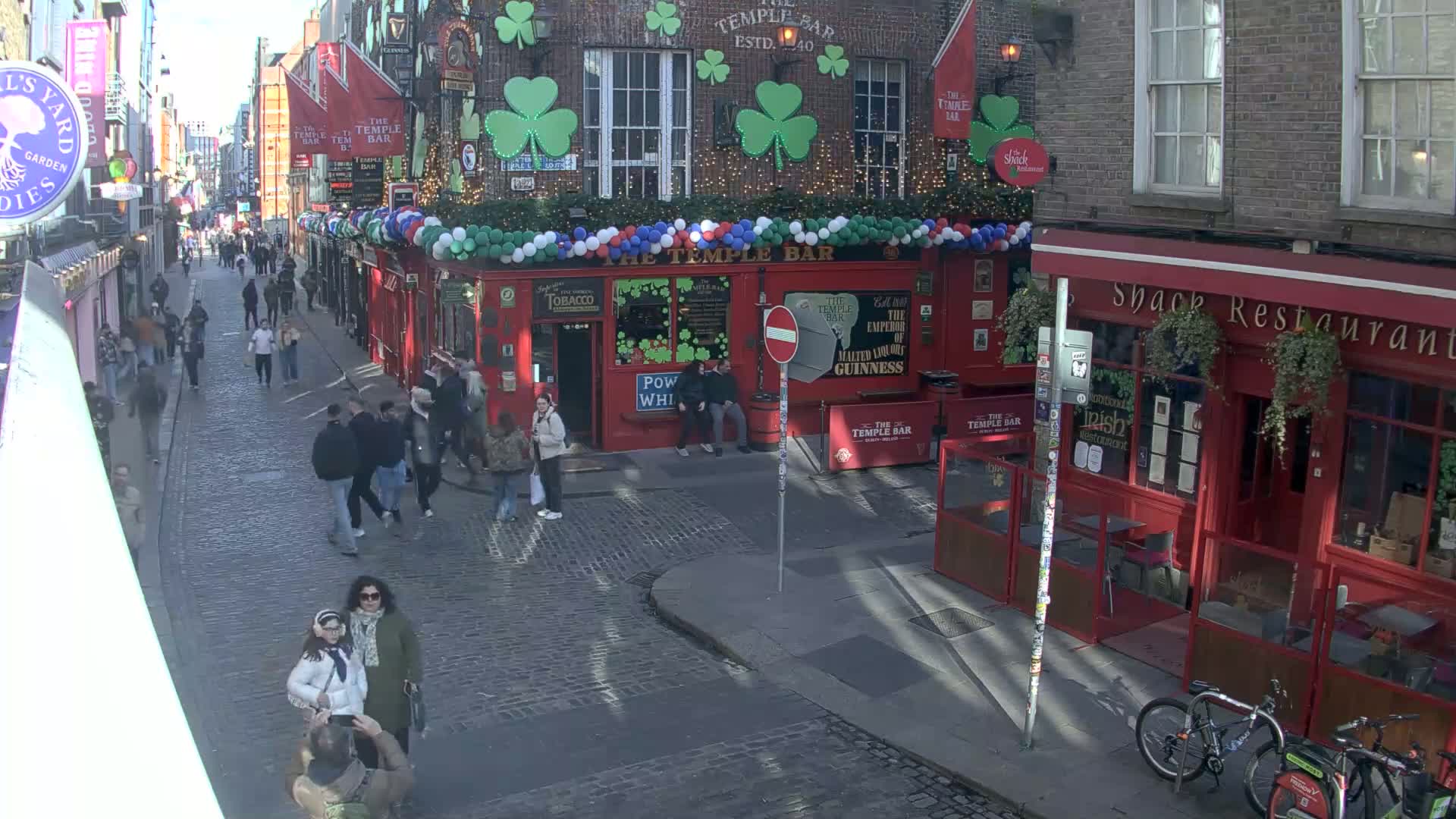 A cobblestone street scene shows many pedestrians walking past red brick buildings, one of which is decorated with shamrocks and balloons, under a sunny sky.