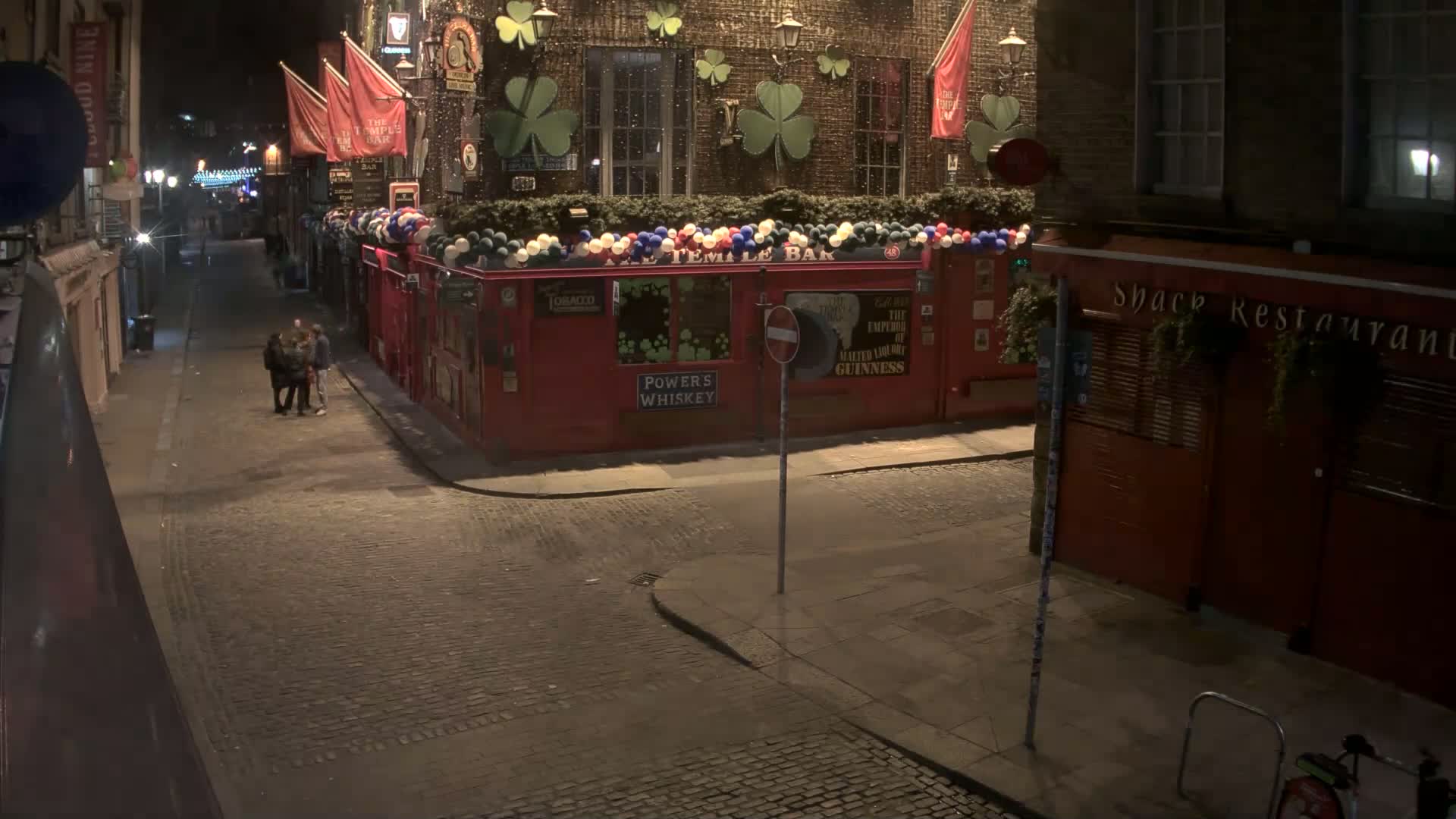 A nighttime street scene shows a cobblestone street with a red pub decorated with shamrocks and balloons, and a small group of people standing nearby in light rain.