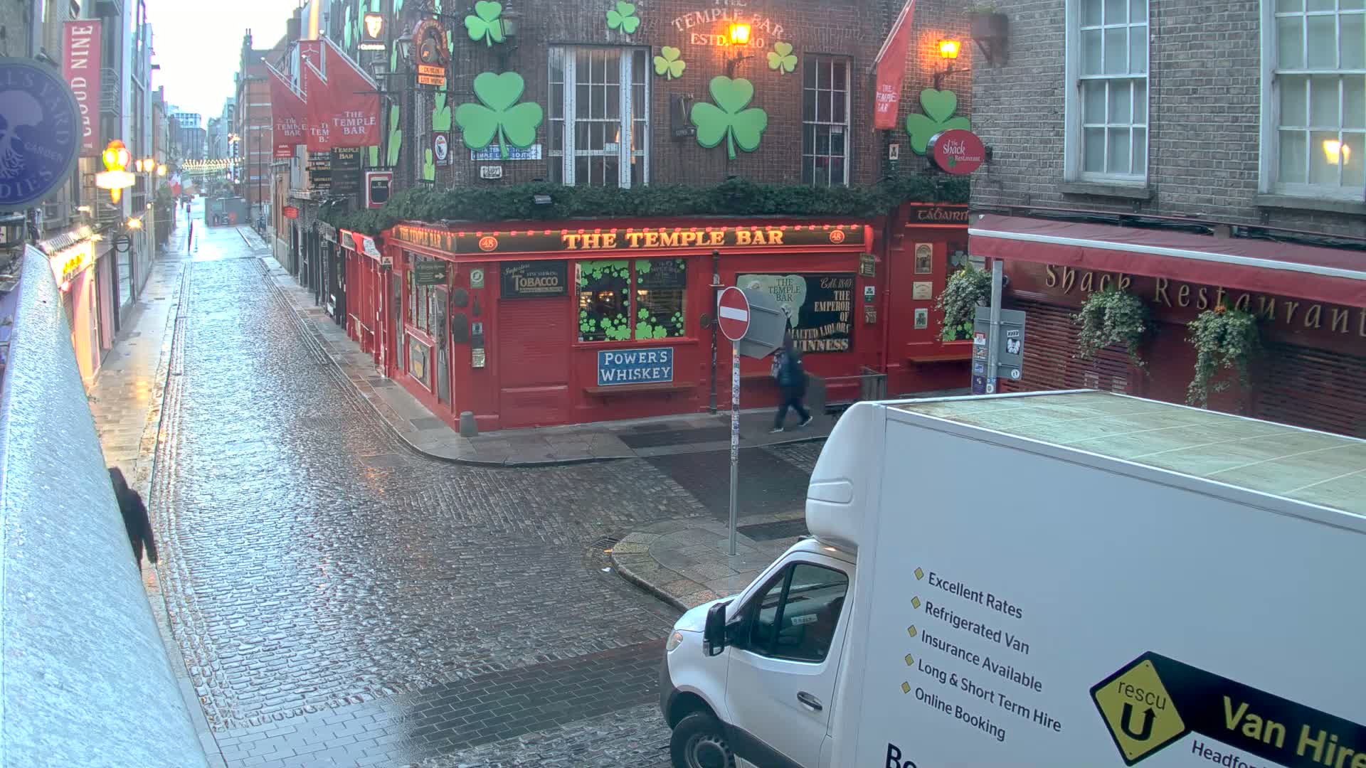 A wet, cobblestone street in a city, featuring a red pub with shamrocks painted on the building, and a white van parked at the corner.