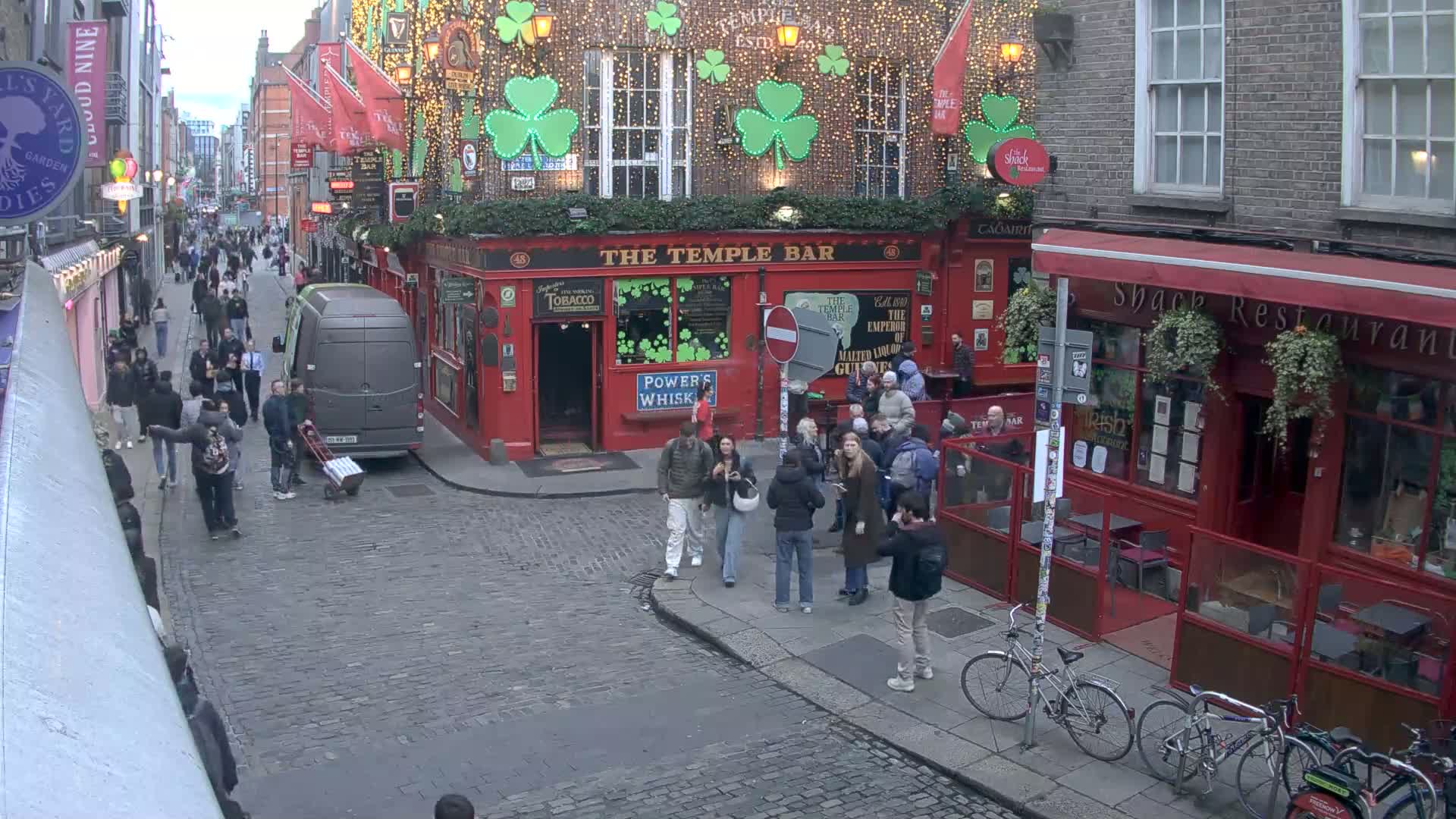 A cobblestone street scene shows a crowd of people walking past a red building with "The Temple Bar" sign and shamrock decorations, under an overcast sky.
