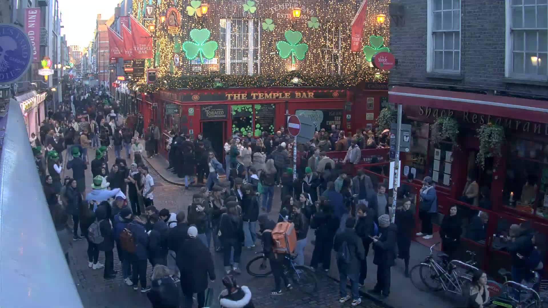 A large crowd of people gathers on a cobblestone street at dusk outside a red brick building decorated with shamrocks and string lights.
