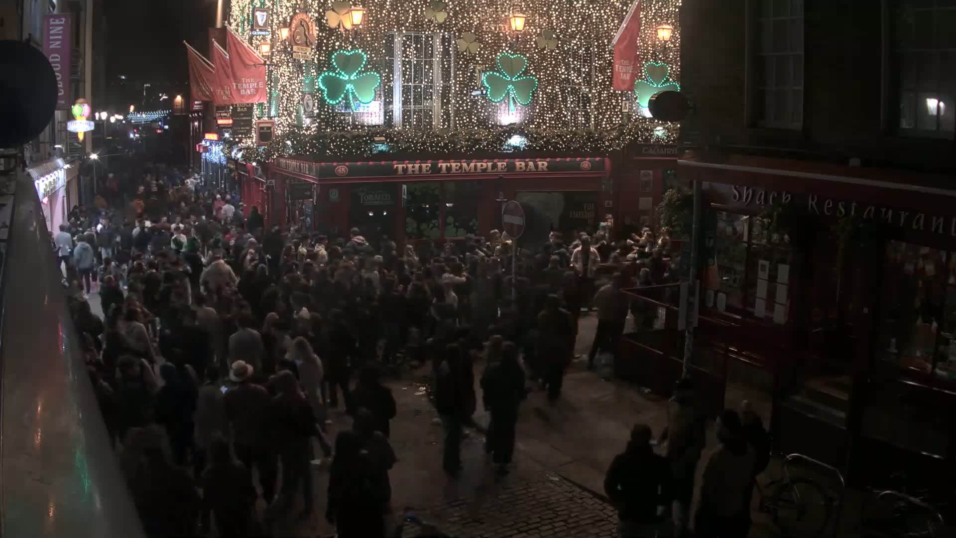 A large crowd of people gathers on a city street at night, under the illuminated facade of a bar decorated with shamrocks and string lights.
