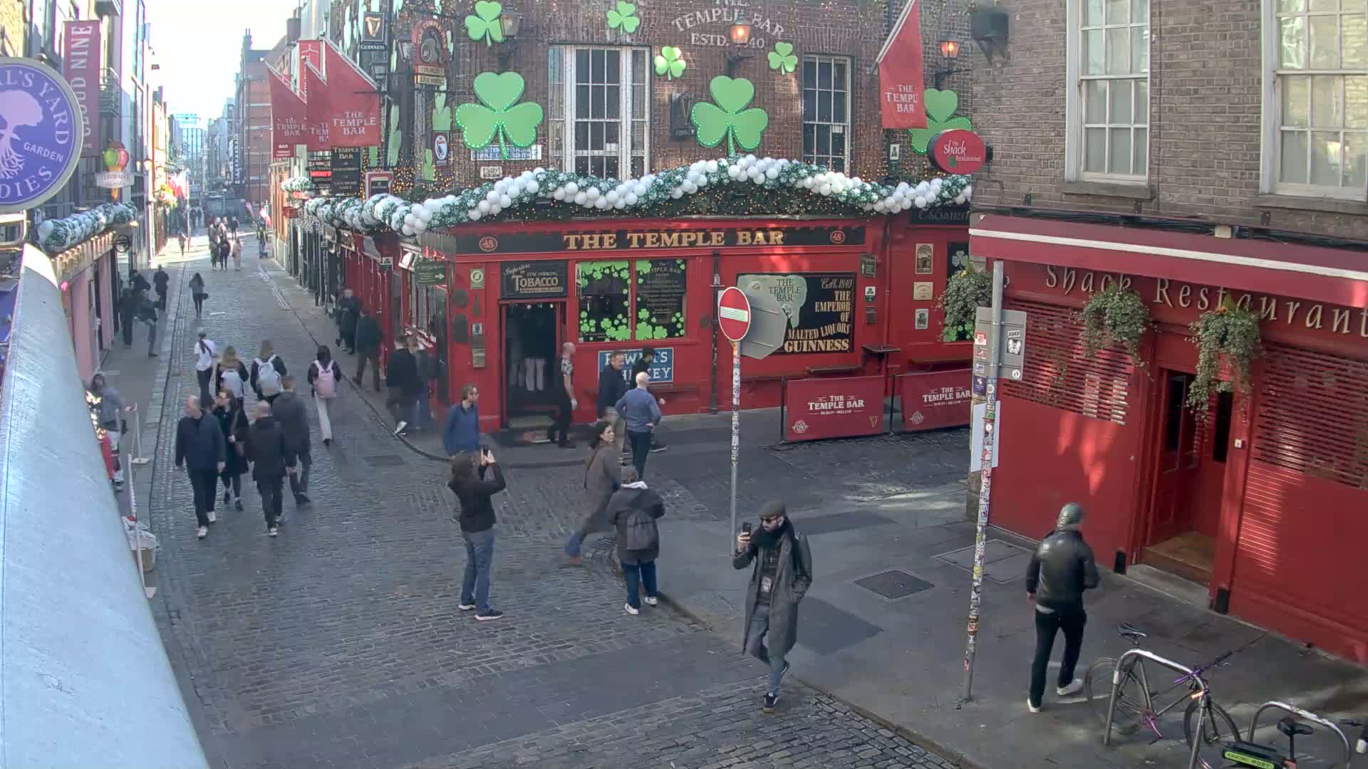A cobblestone street scene shows a crowd of people walking past red brick buildings decorated with shamrocks and white lights, under a bright, sunny sky.