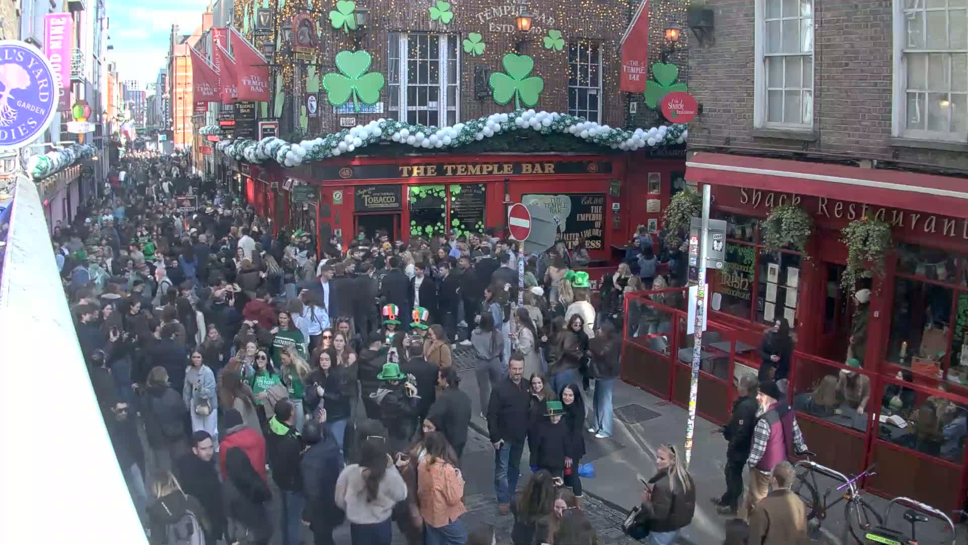 A crowded street scene on a sunny day, with many people gathered outside a pub decorated with shamrocks.