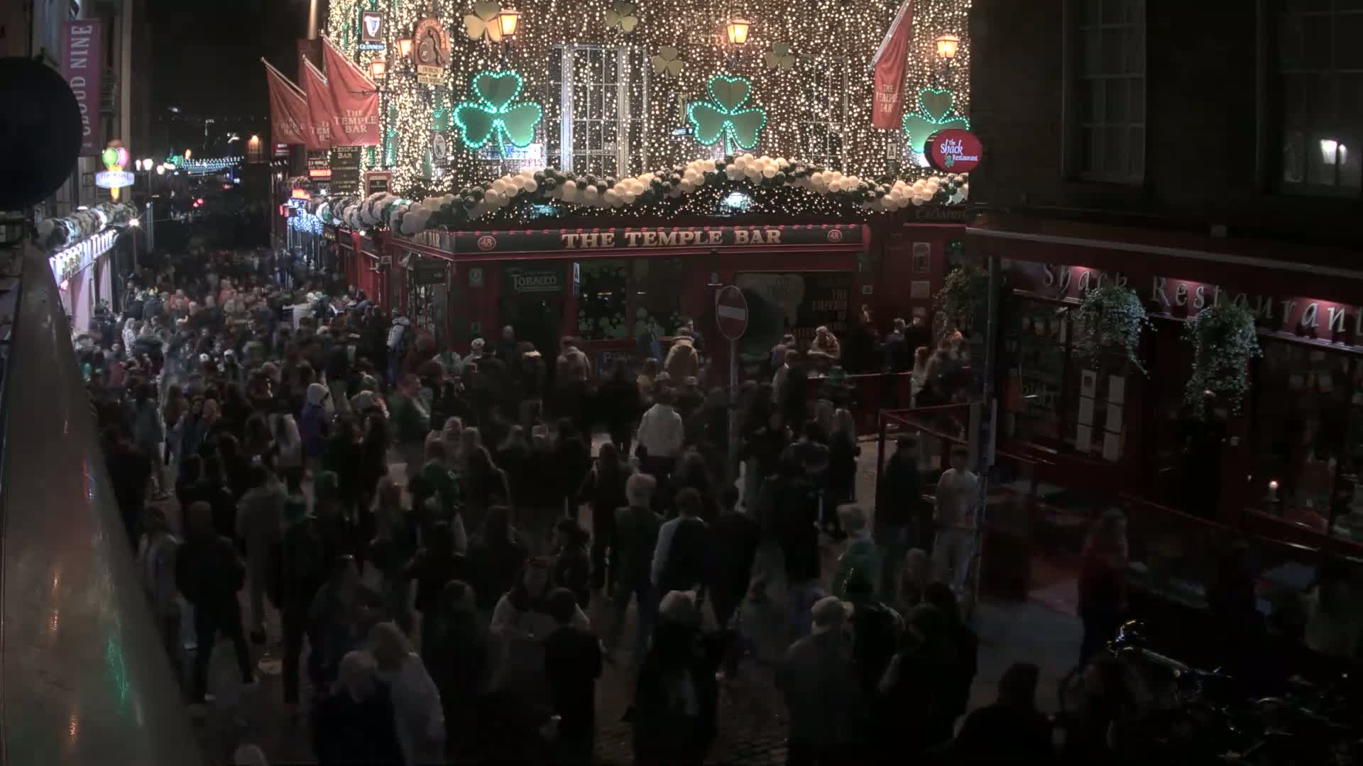 A large crowd of people gathers on a nighttime street outside a brightly lit pub decorated with shamrocks and lights.