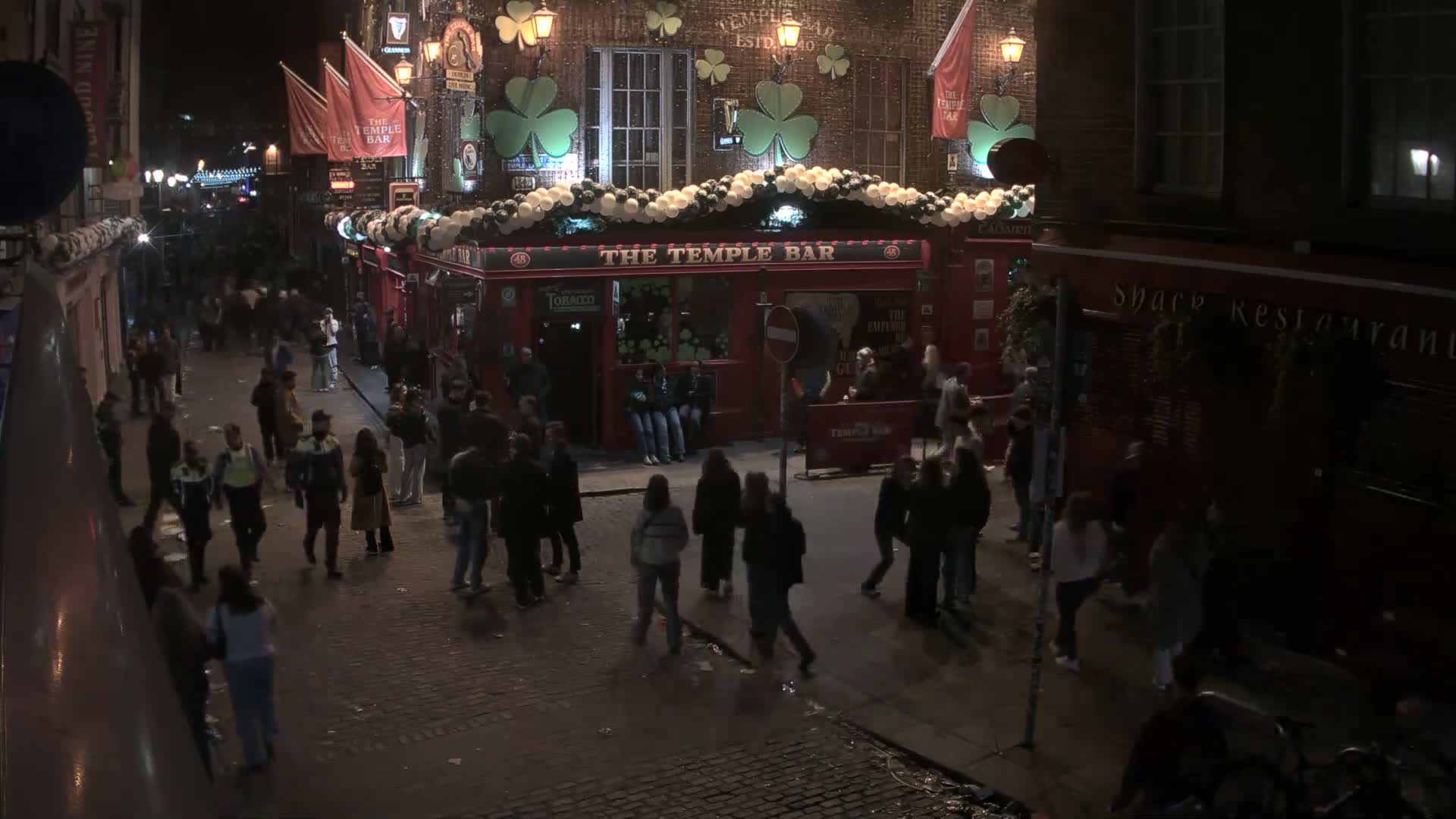 A nighttime street scene shows a crowd of people gathered outside a red brick pub decorated with shamrocks and white balloons under lightly falling snow.