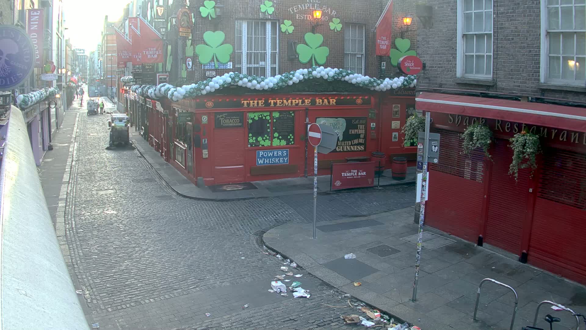 A cobblestone street, littered with trash, shows a red building decorated with shamrocks and white balloons, next to another red building with its shutters closed, under a mostly clear, bright sky.