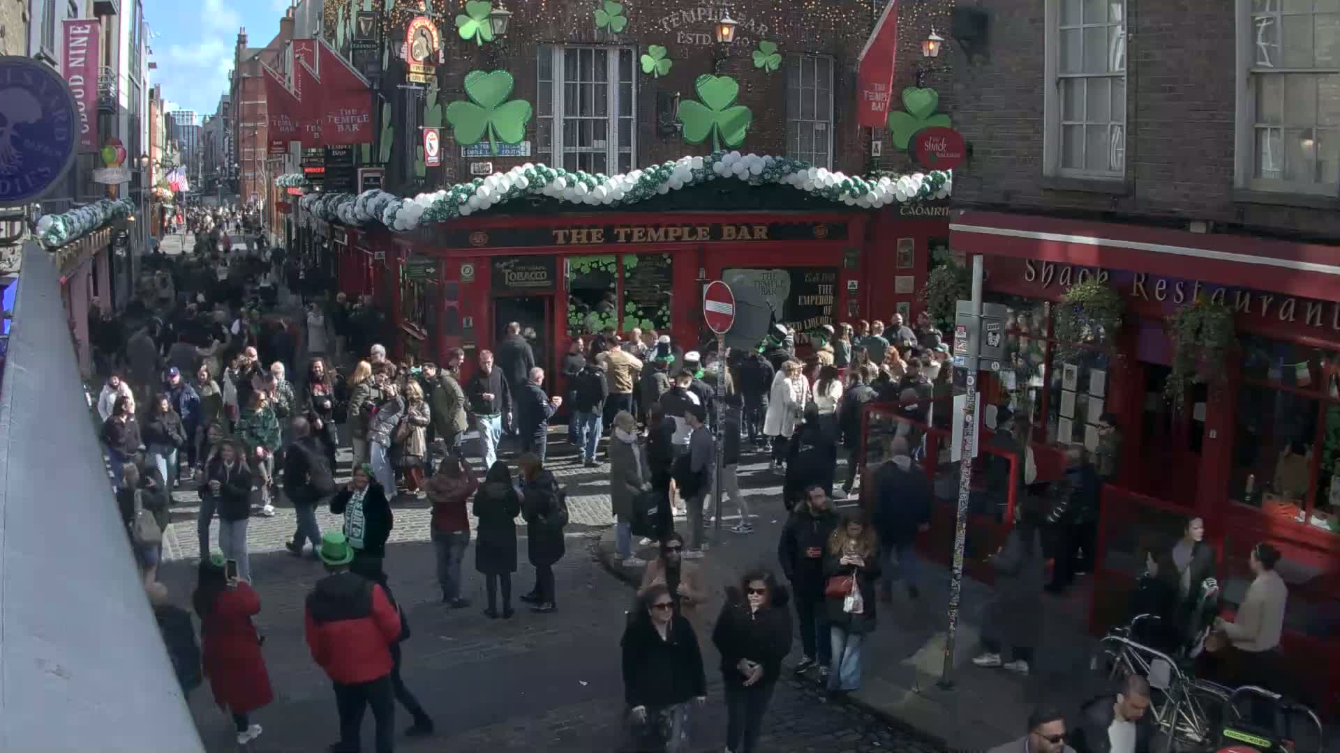 A crowded street scene on a sunny day, filled with pedestrians near a red building decorated with shamrocks and balloons.