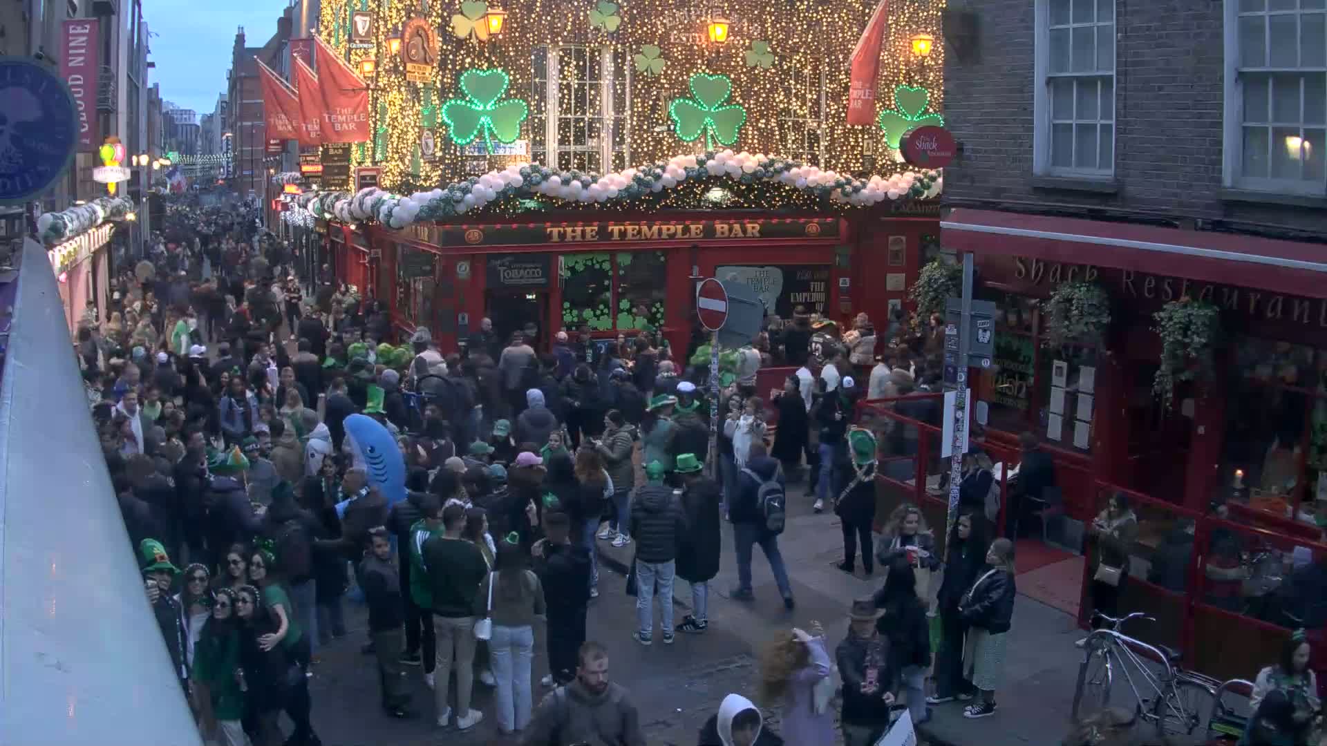 A large crowd of people, many wearing green, gathers on a city street at dusk, in front of a brightly lit pub decorated with shamrocks and lights.