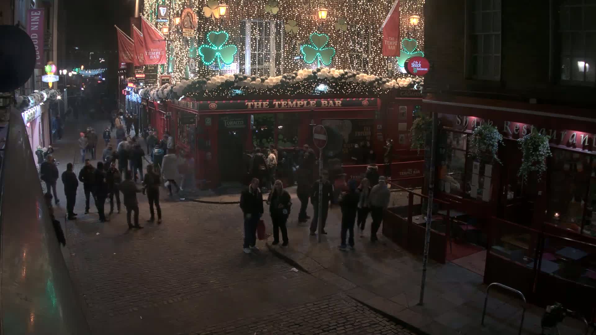A nighttime street scene shows a crowd of people gathered outside a brightly lit pub decorated with shamrocks and string lights.