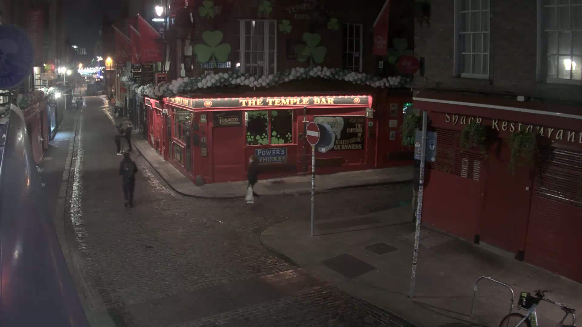 A wet cobblestone street at night, with a few pedestrians walking past a red pub and restaurant.