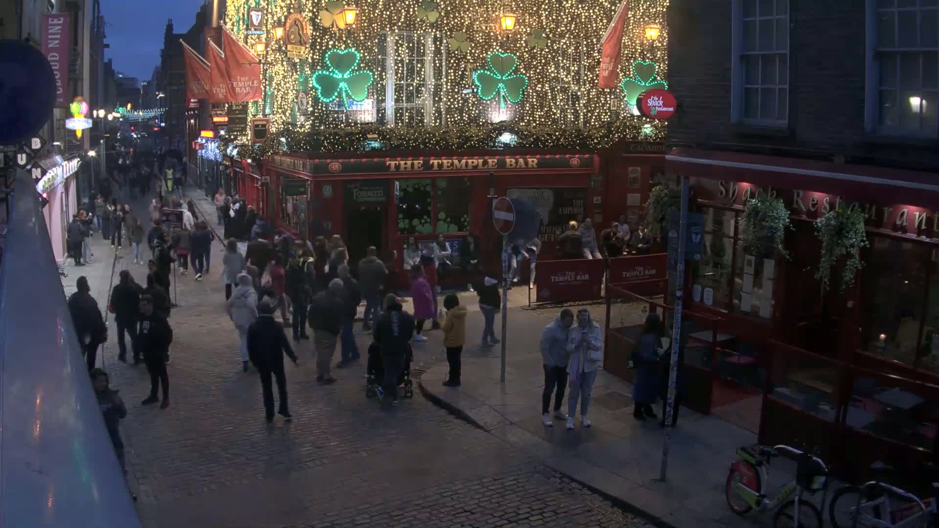 A bustling, nighttime street scene features a crowd of people gathered outside a brightly lit pub decorated with shamrocks and string lights.