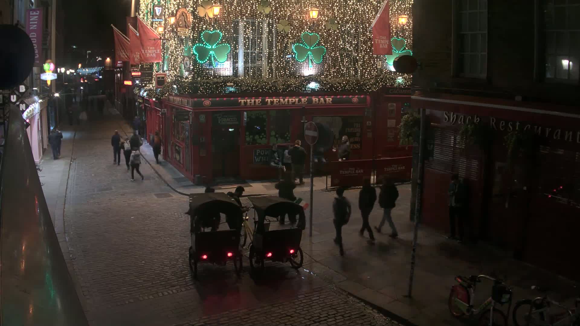 At night, a cobblestone street is illuminated by the lights of a brightly decorated pub and surrounding buildings, with several pedestrians and two rickshaws present.