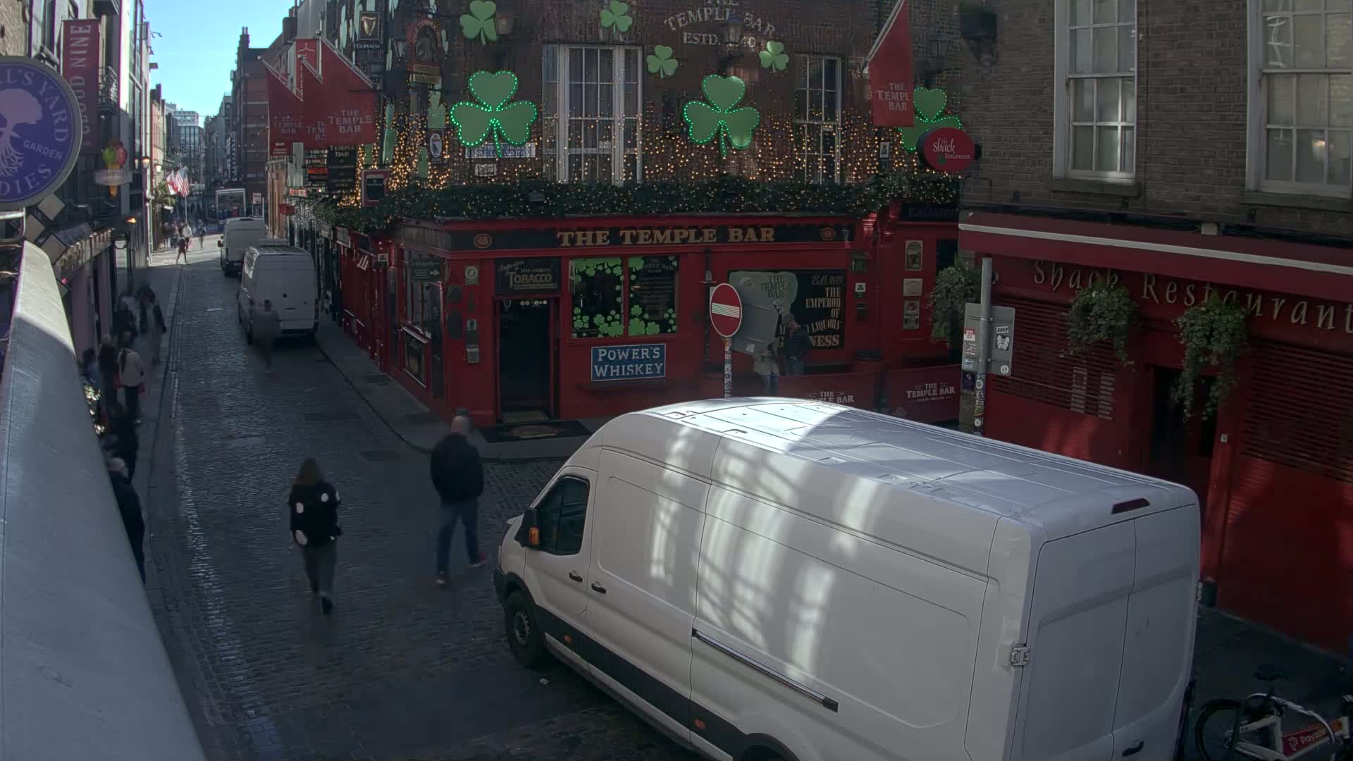 A cobblestone street, under a sunny sky, shows pedestrians walking past a red brick building decorated with shamrocks and string lights, and parked vans.