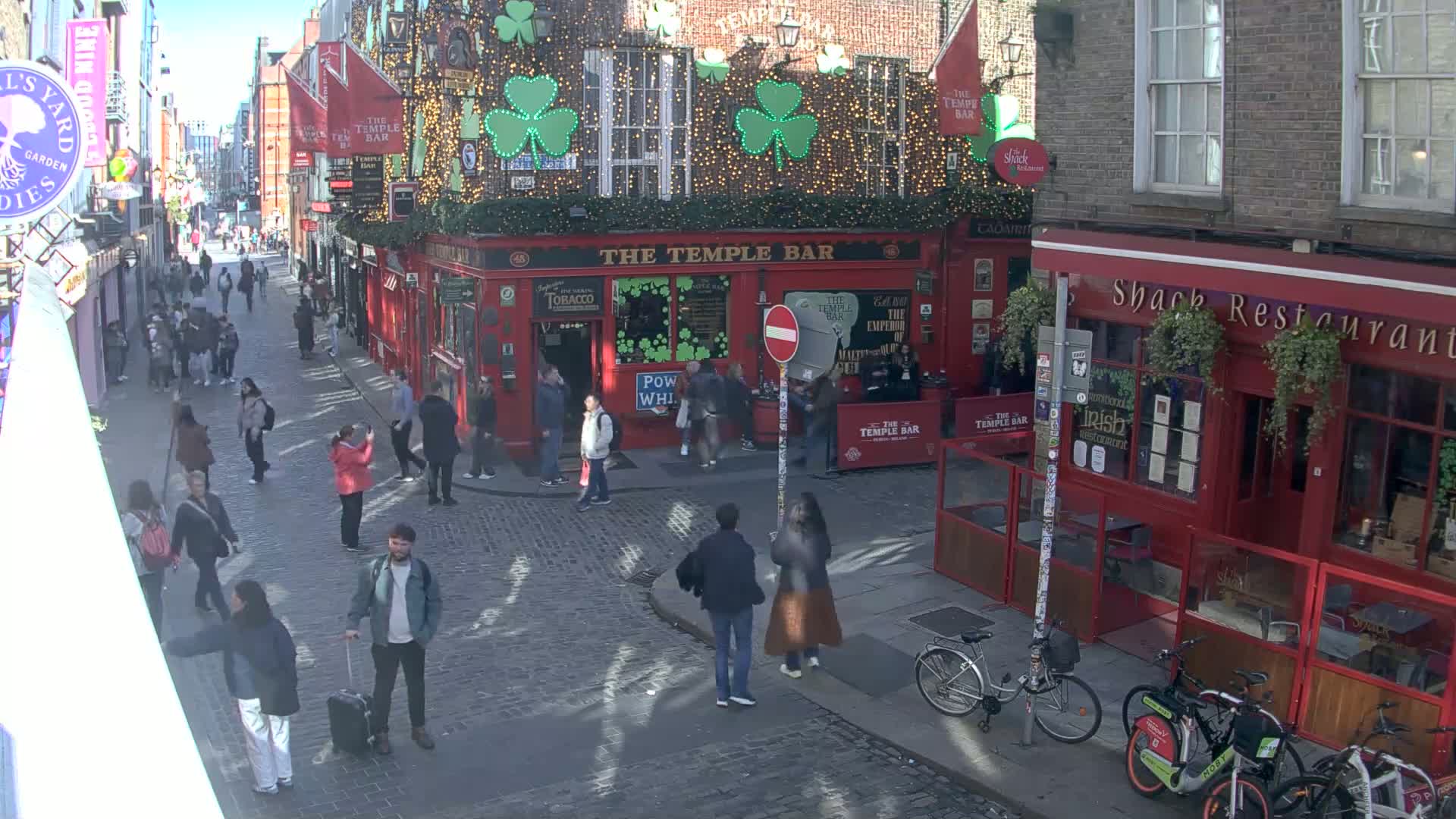 A cobblestone street scene shows numerous pedestrians, some carrying luggage, walking past red brick buildings, including a pub decorated with shamrocks and lights, under a sunny sky.