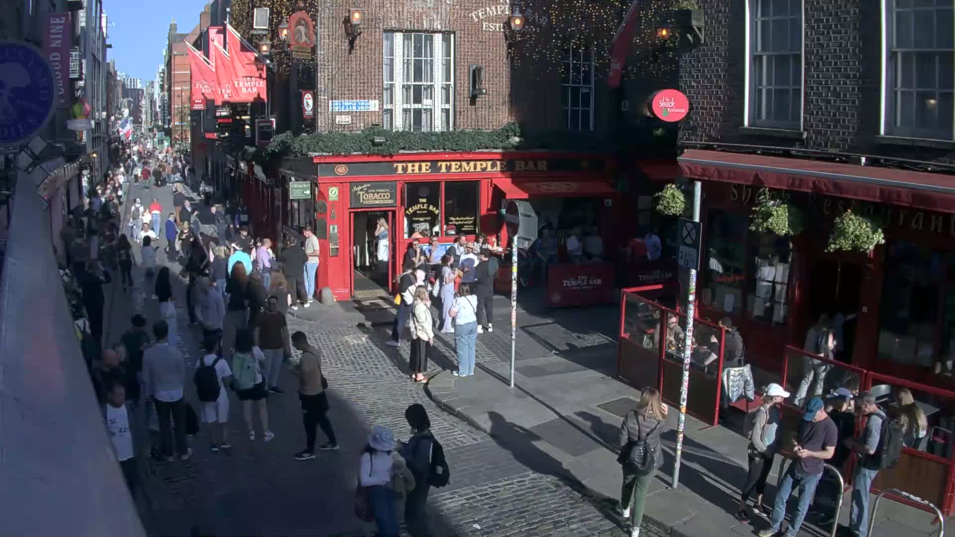 A sunny day on a cobblestone street, crowded with people walking past red brick buildings and outdoor seating areas of pubs and restaurants.