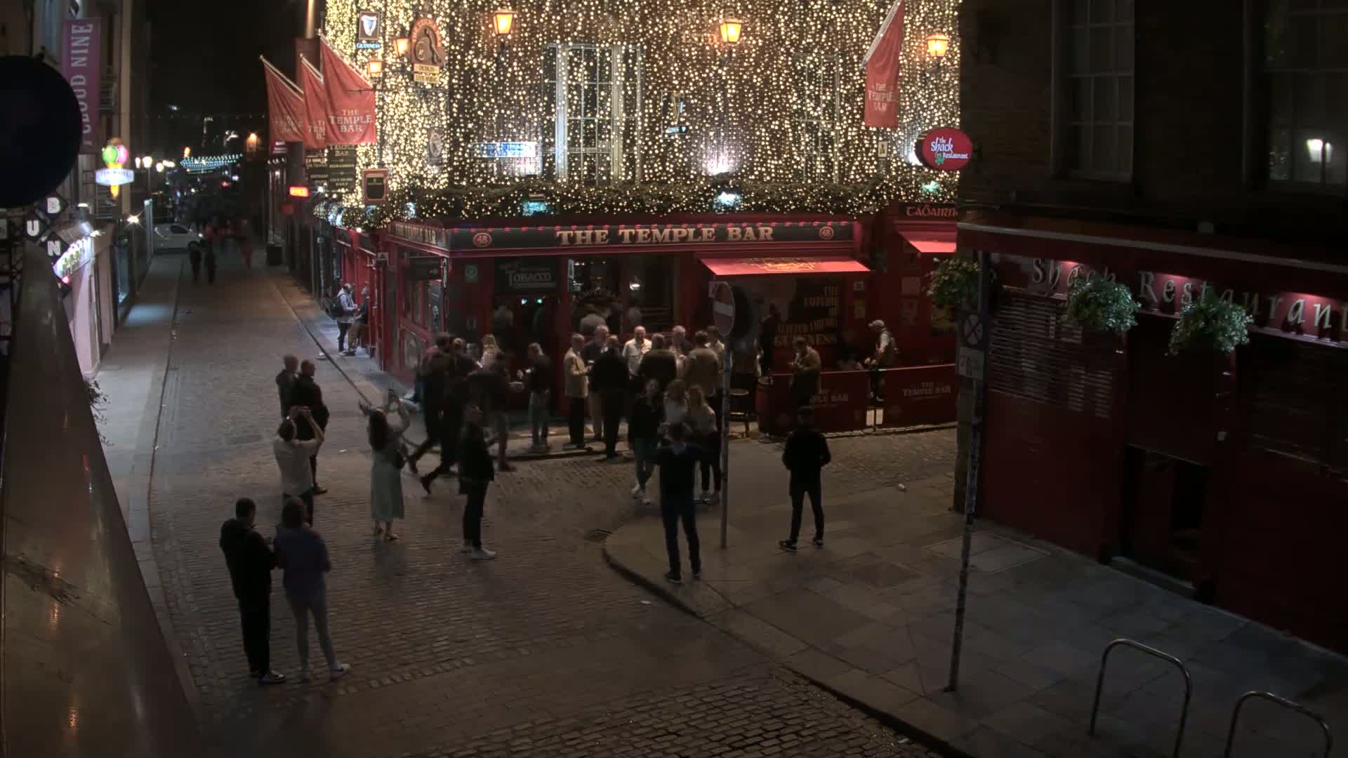 A nighttime, cobblestone street scene shows a crowd of people gathered outside a brightly lit pub adorned with string lights, and other people walking down the street.