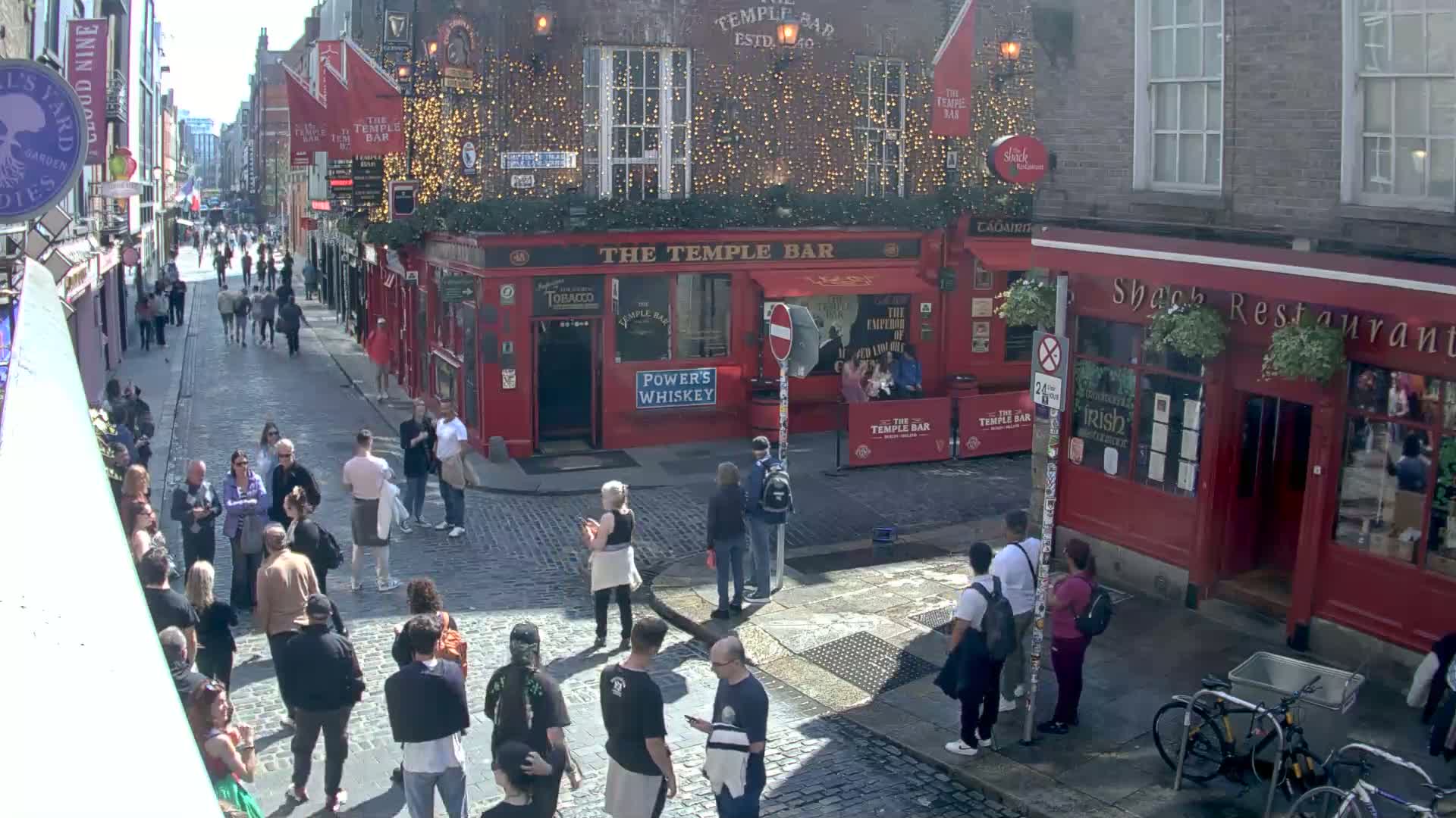 A sunny cobblestone street scene shows numerous people walking past red brick buildings, including a pub called "The Temple Bar".