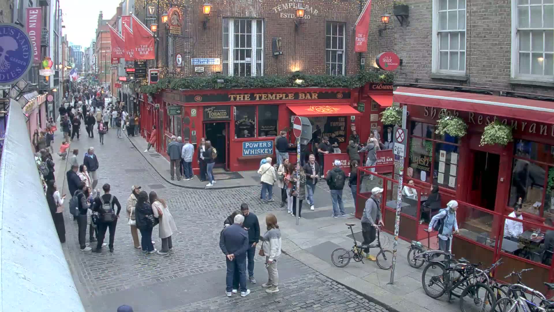 A cobblestone street in overcast weather is crowded with pedestrians and cyclists, lined with red brick buildings including a prominent pub.