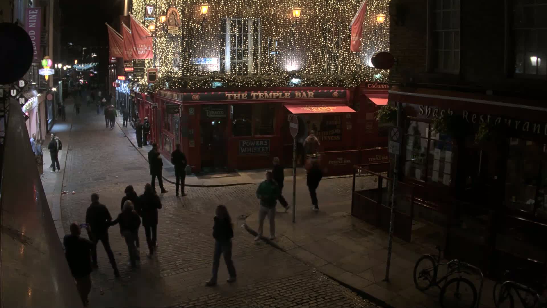 A nighttime street scene shows people walking past a brightly lit pub decorated with string lights, and a darker restaurant next door, with bicycles parked on the sidewalk.