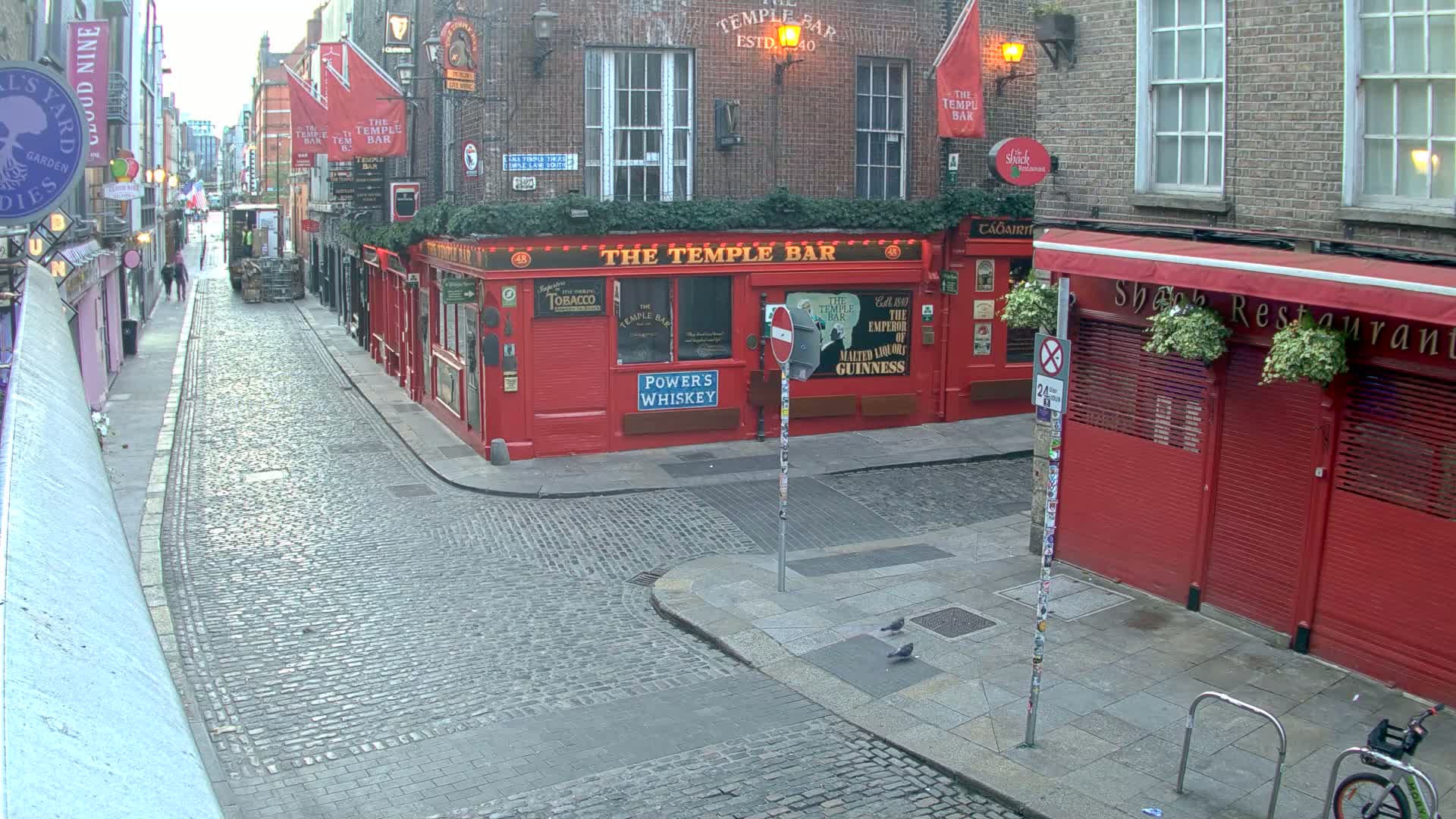 A cobblestone street, under an overcast sky, features red-brick buildings, including a prominent red pub, and a few pedestrians.