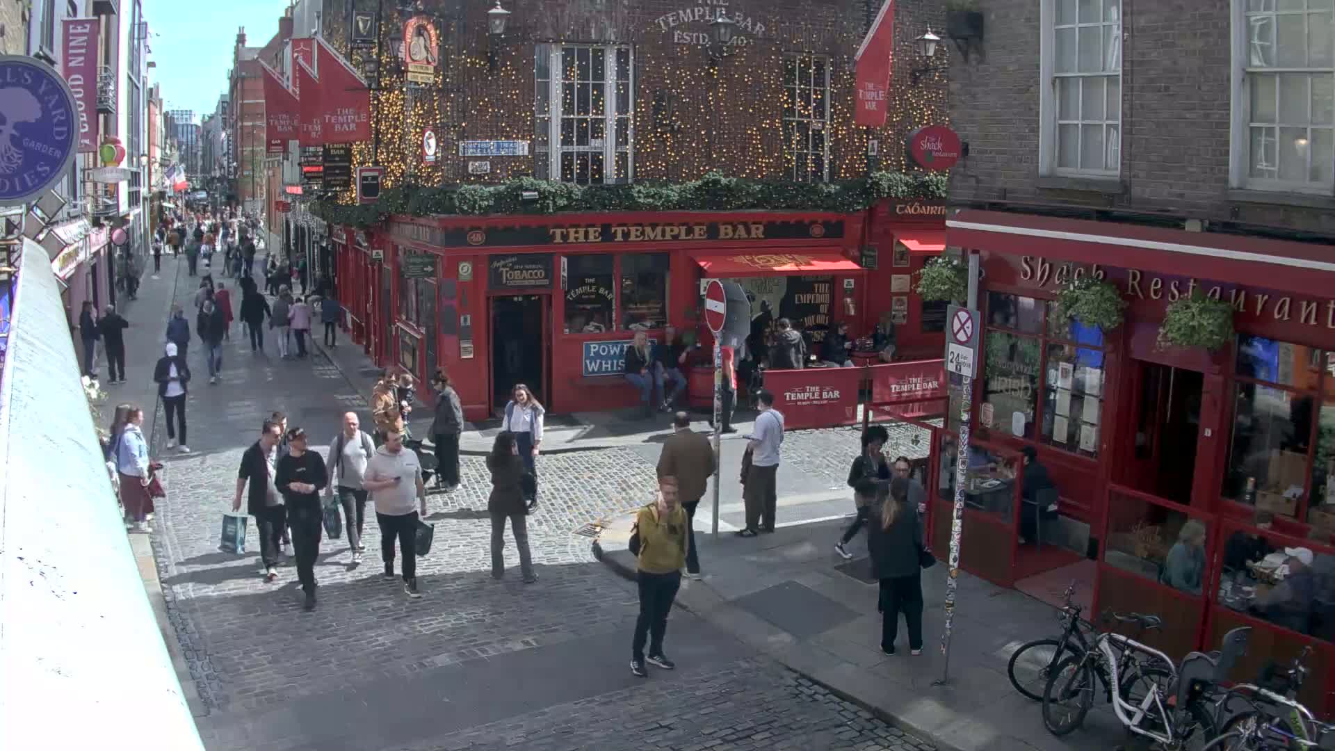 A bustling cobblestone street features pedestrians walking past red brick buildings, one of which is a pub with outdoor seating, under a sunny sky.