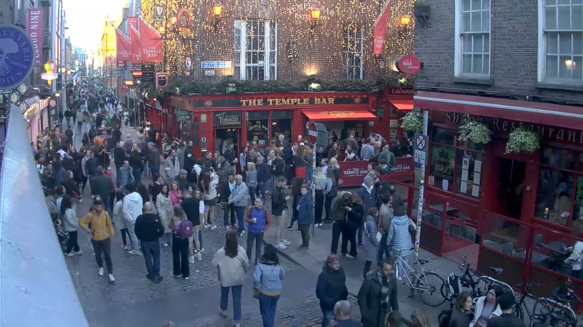 A large crowd of people gathers in a cobblestone street at dusk, near a red brick building decorated with string lights.