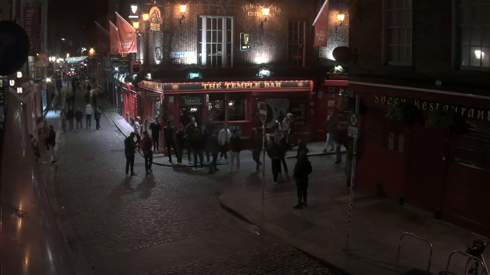 A cobblestone street at night, lined with red buildings and populated by groups of people near a brightly lit pub.