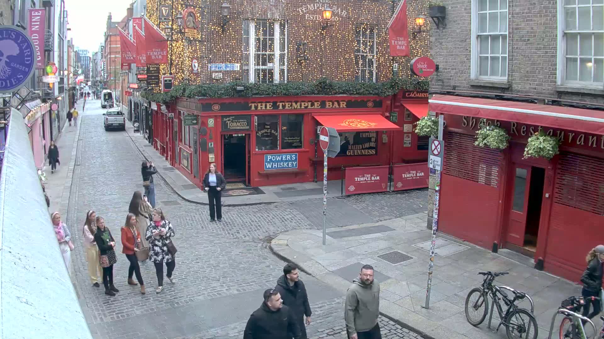 A cobblestone street scene shows pedestrians walking past a red building with string lights and a red awning, along with bicycles parked nearby under an overcast sky.